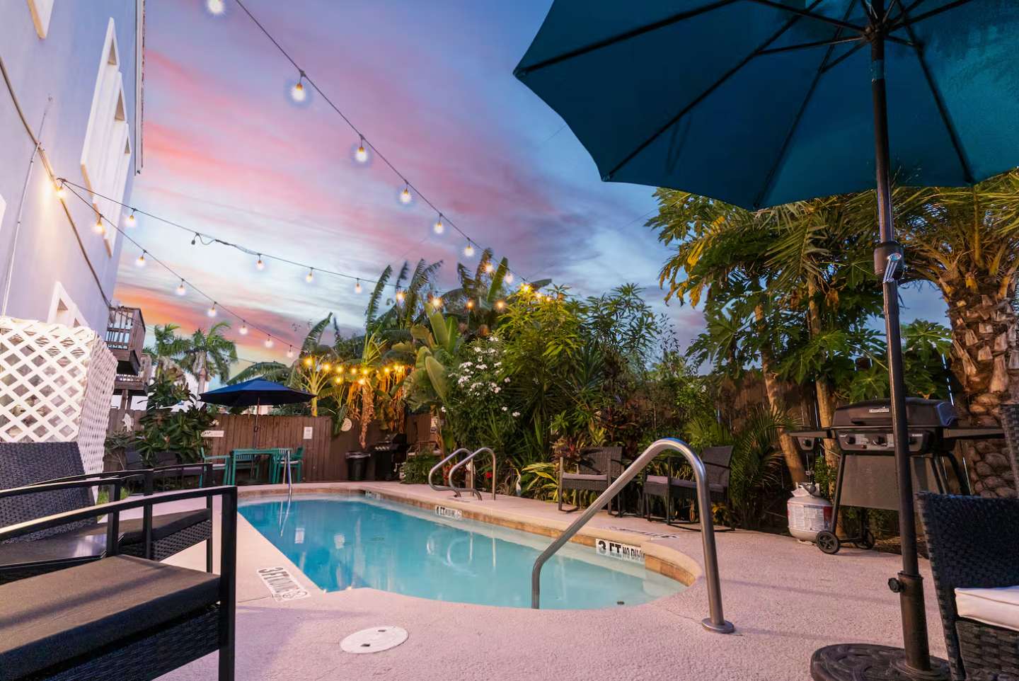 Inviting outdoor pool area at dusk, illuminated by string lights, surrounded by lush tropical foliage and palm trees, with a blue umbrella, various seating, and a grill nearby.