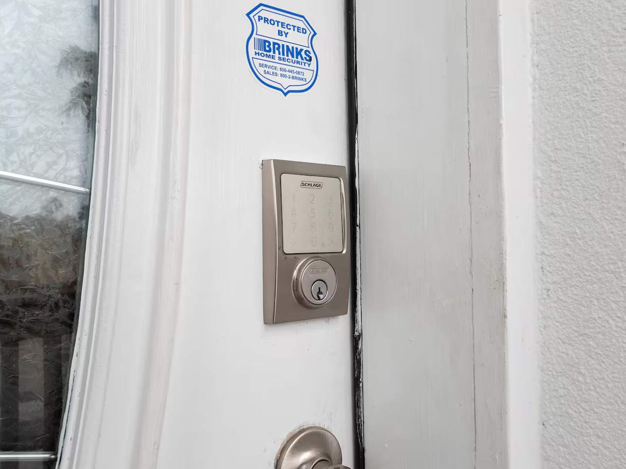 A close-up view of a white exterior door featuring a modern silver keypad smart lock and a traditional deadbolt, with a "Protected by Brinks Home Security" sticker visible. The door has a decorative glass insert.