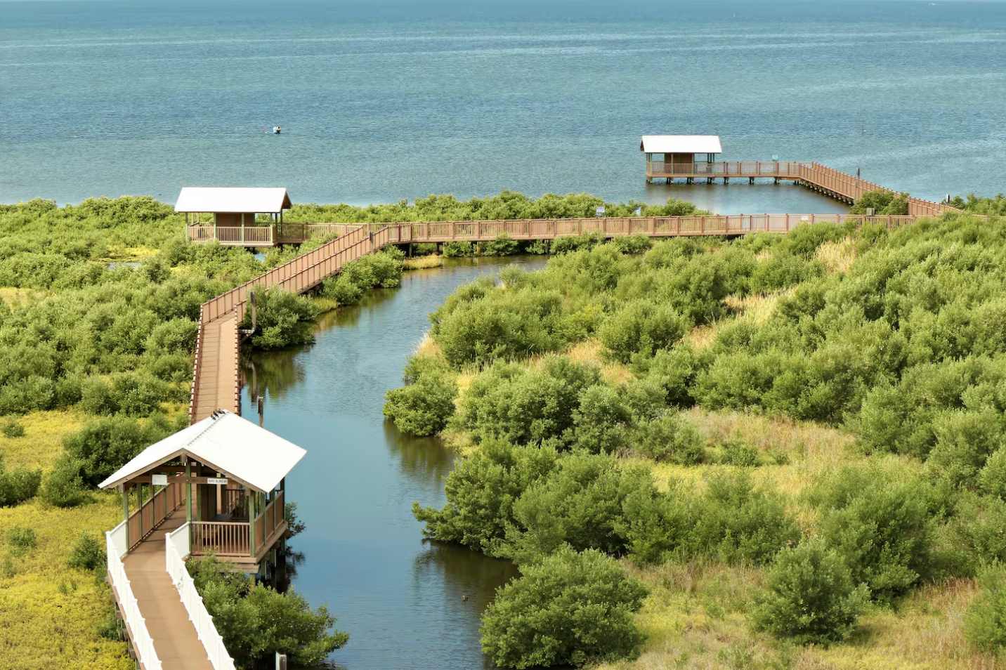 Elevated view of a winding wooden boardwalk traversing lush green marshland, connecting small covered gazebos over water. A large body of water is visible in the distance.