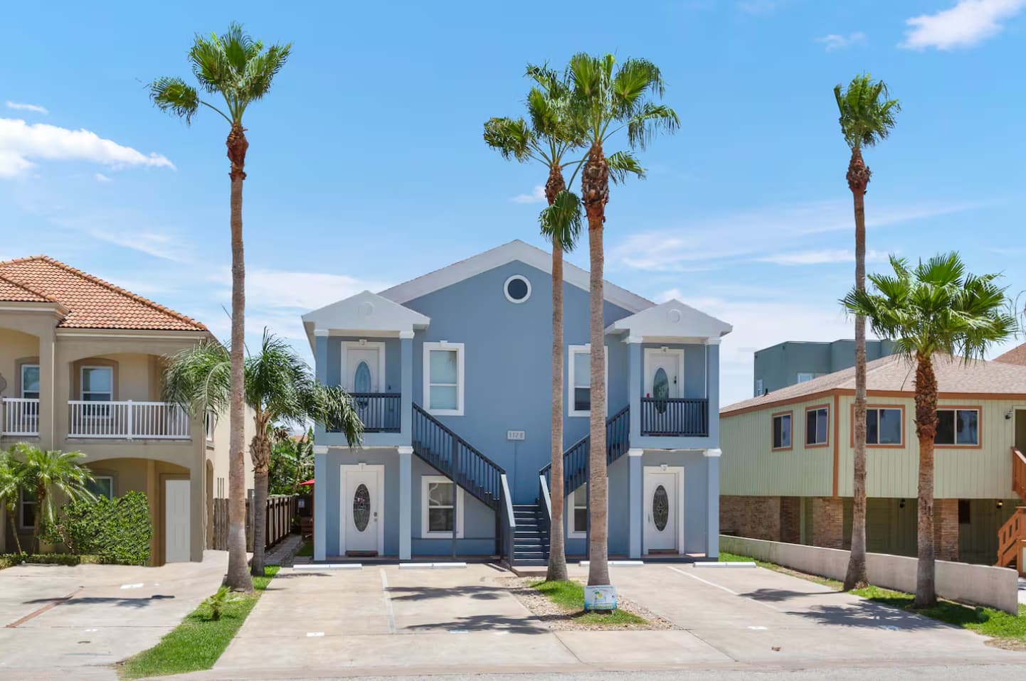 The front exterior of a two-story blue apartment building with white trim and multiple tall palm trees, featuring two external staircases leading to upper-level balconies. The scene is set under a bright blue sky with scattered white clouds.