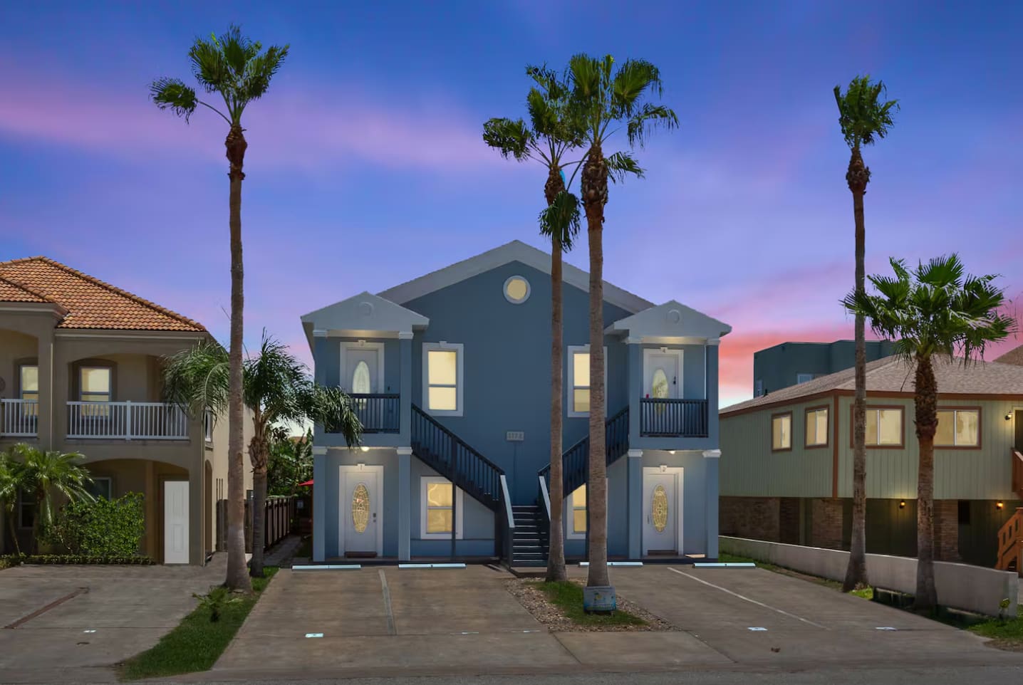 The front exterior of a two-story blue apartment building with white trim and multiple palm trees, featuring two external staircases leading to upper-level balconies. The sky is a dramatic mix of pink, purple, and blue at dusk.