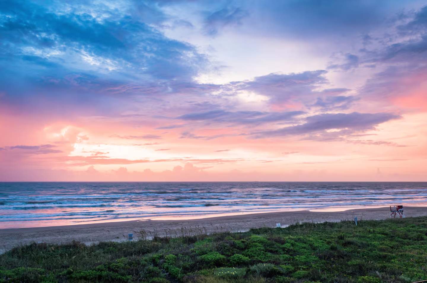 Vibrant sunset or sunrise over a beach, casting pink, orange, and blue hues across the sky and reflecting on the ocean waves. Green dune vegetation and a lifeguard chair are visible in the foreground.