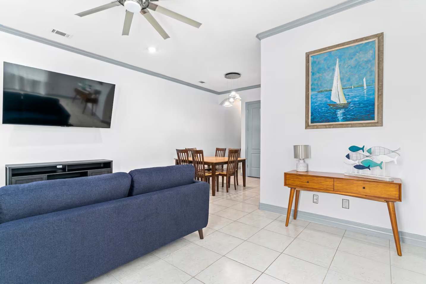 Open concept living and dining area with a navy blue sofa facing a wall-mounted TV. A wooden dining table and chairs are visible in the background, all on white tile flooring.