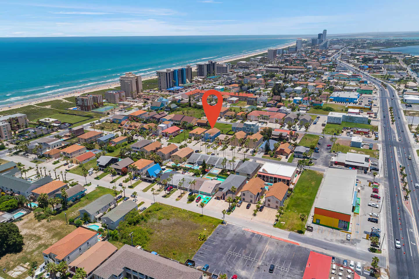 An aerial view showcases the property's location within a coastal town, marked by a red pin, with the ocean and a city skyline visible in the background. The surrounding area features residential homes and commercial buildings.