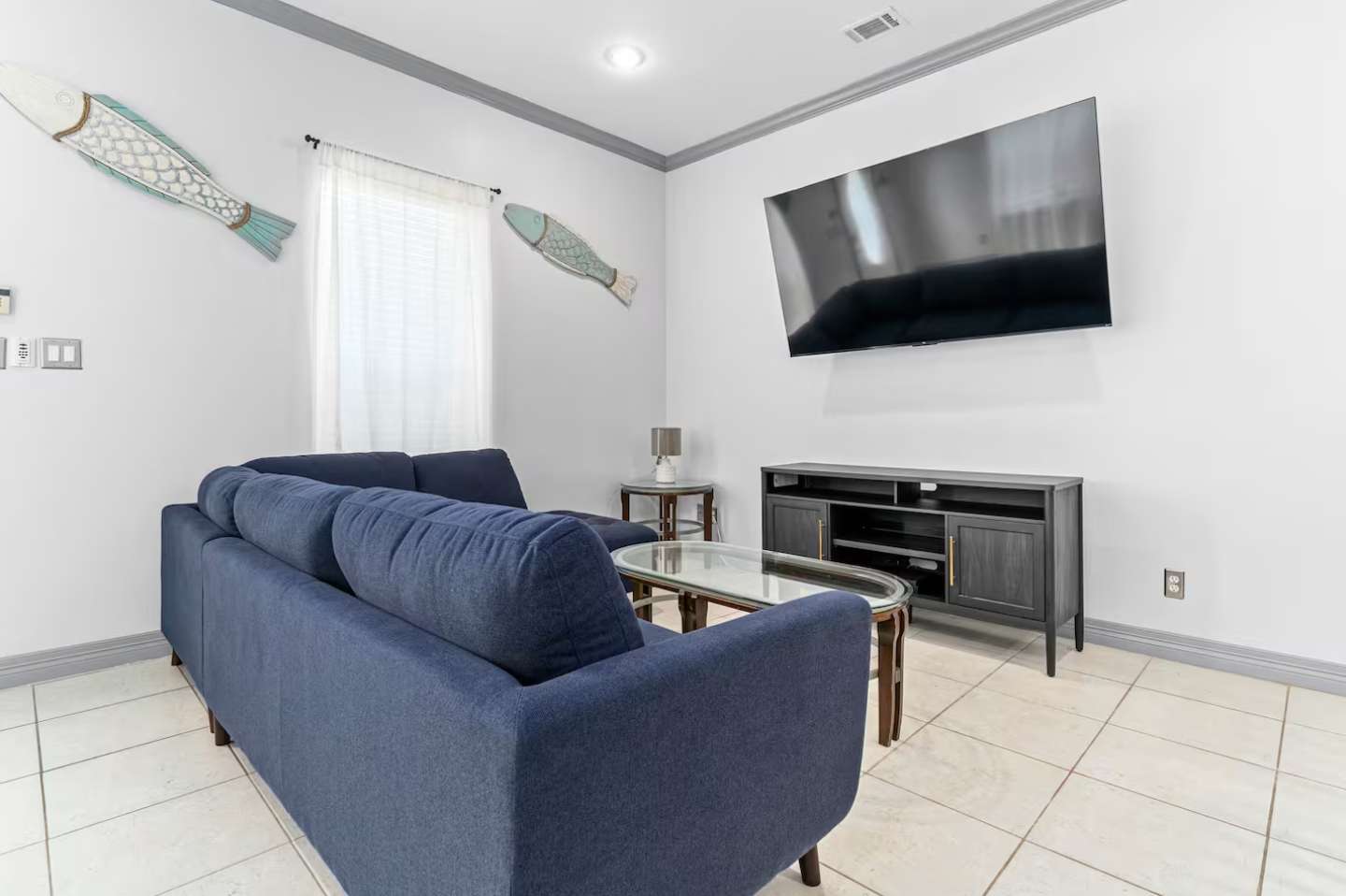 Cozy living room with a navy blue sofa, a glass-top coffee table, and a large wall-mounted TV. Decorative fish adorn the white walls, and sheer curtains cover the windows.