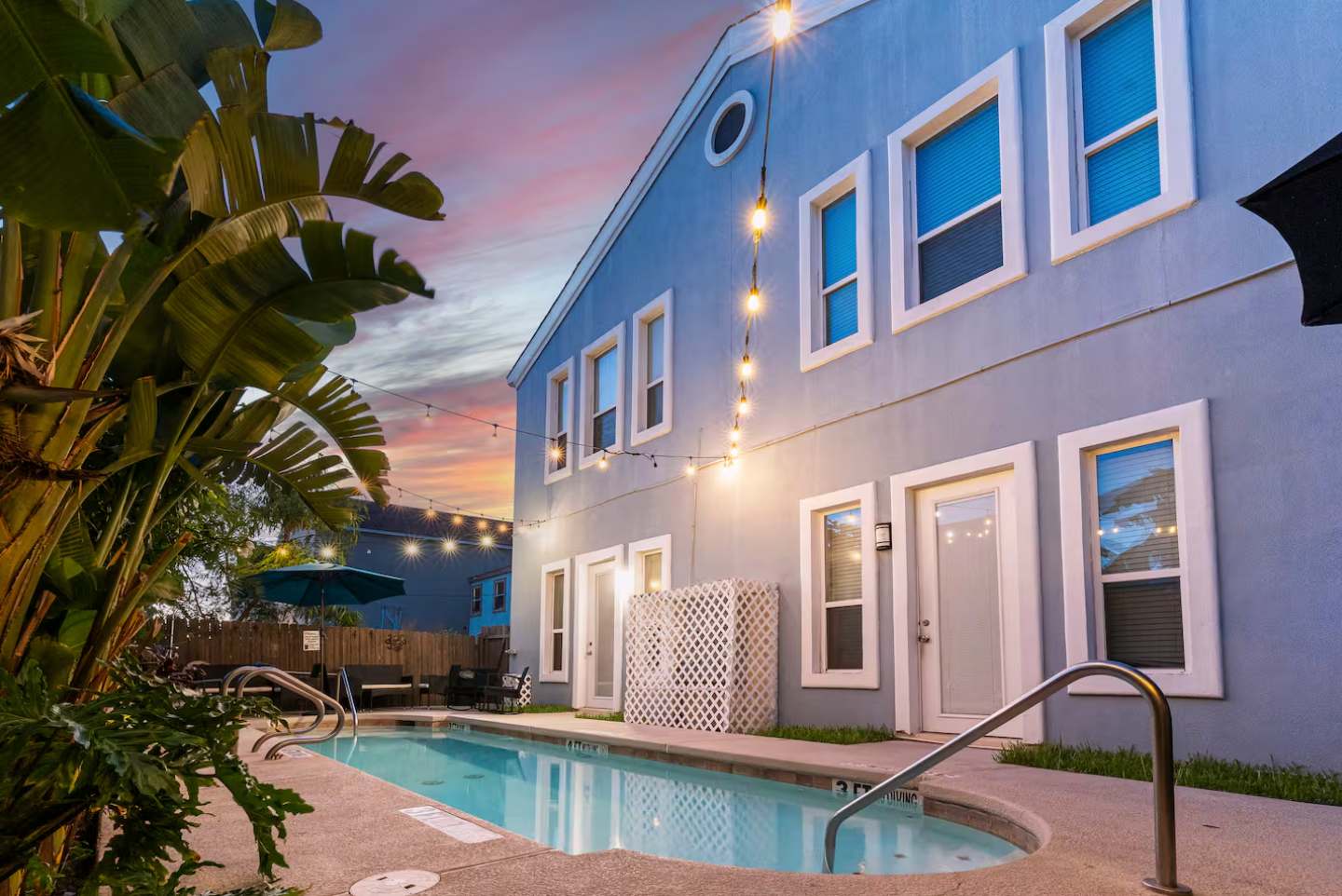 Rear exterior of a light blue two-story building with multiple windows and doors, overlooking a swimming pool. String lights are strung across the yard, and tropical foliage is visible.