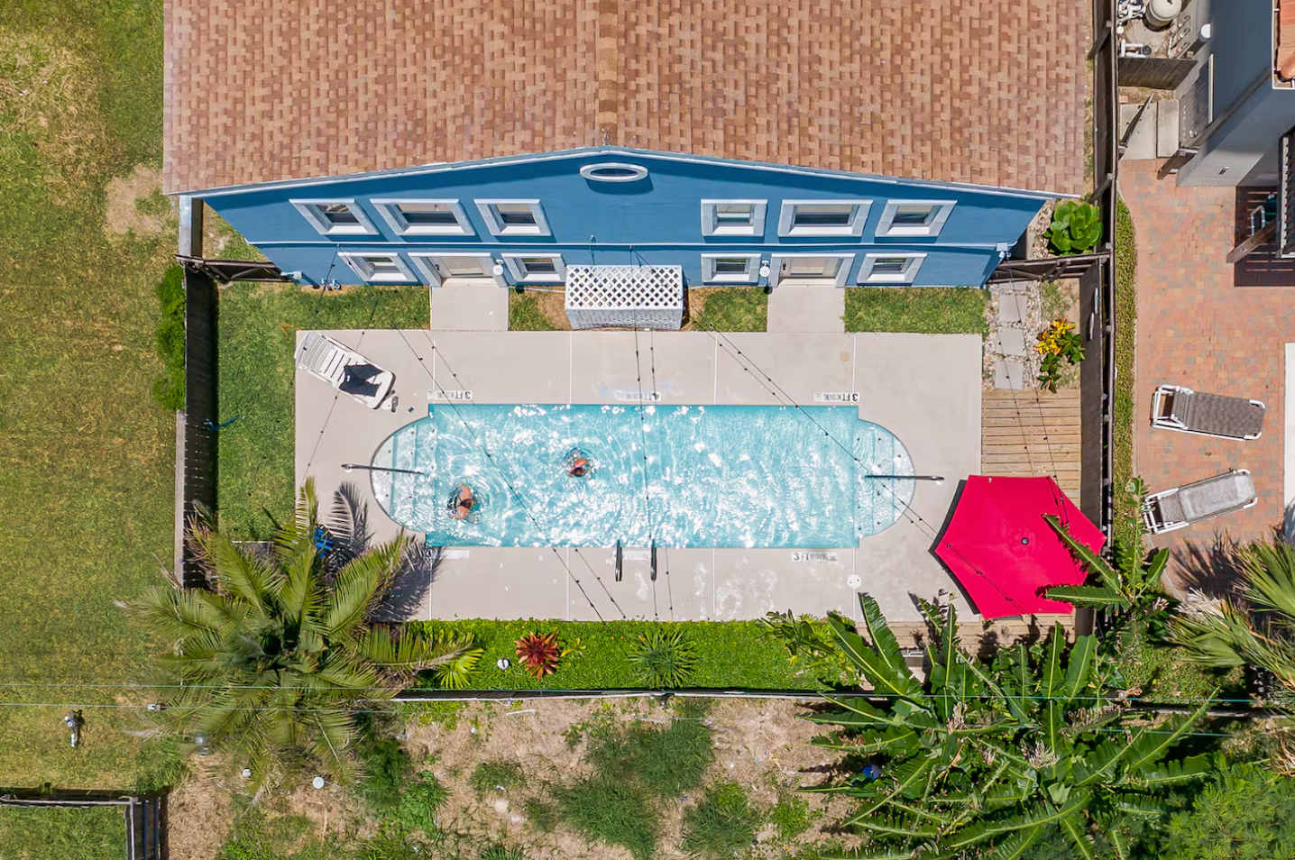 Overhead drone shot revealing a long rectangular swimming pool with two swimmers, surrounded by a concrete patio, lush green landscaping, and a blue multi-story building with a reddish-brown roof.