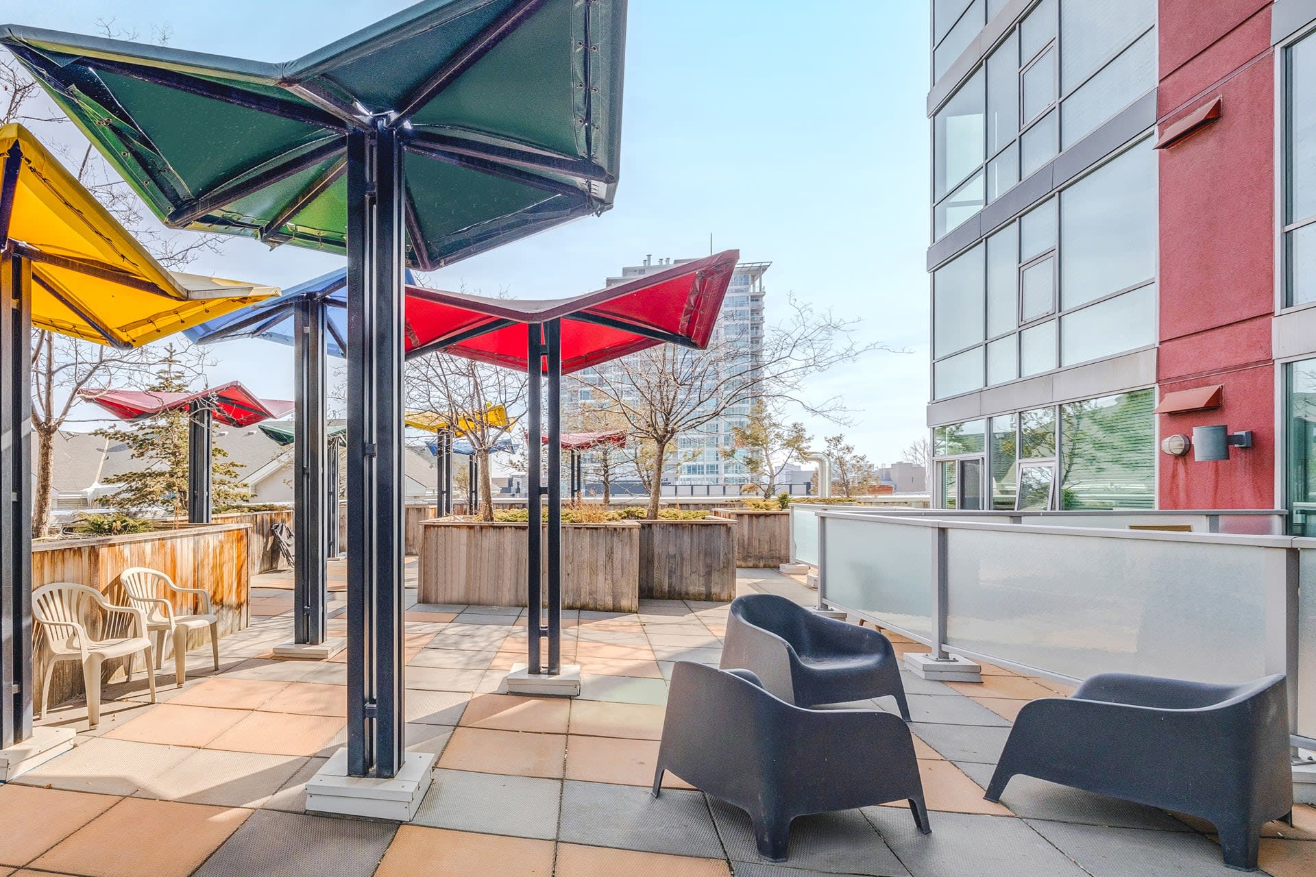 An outdoor common patio area features colorful, star-shaped shade structures over tiled flooring, with dark modern chairs and wooden planters, offering a view of surrounding buildings.
