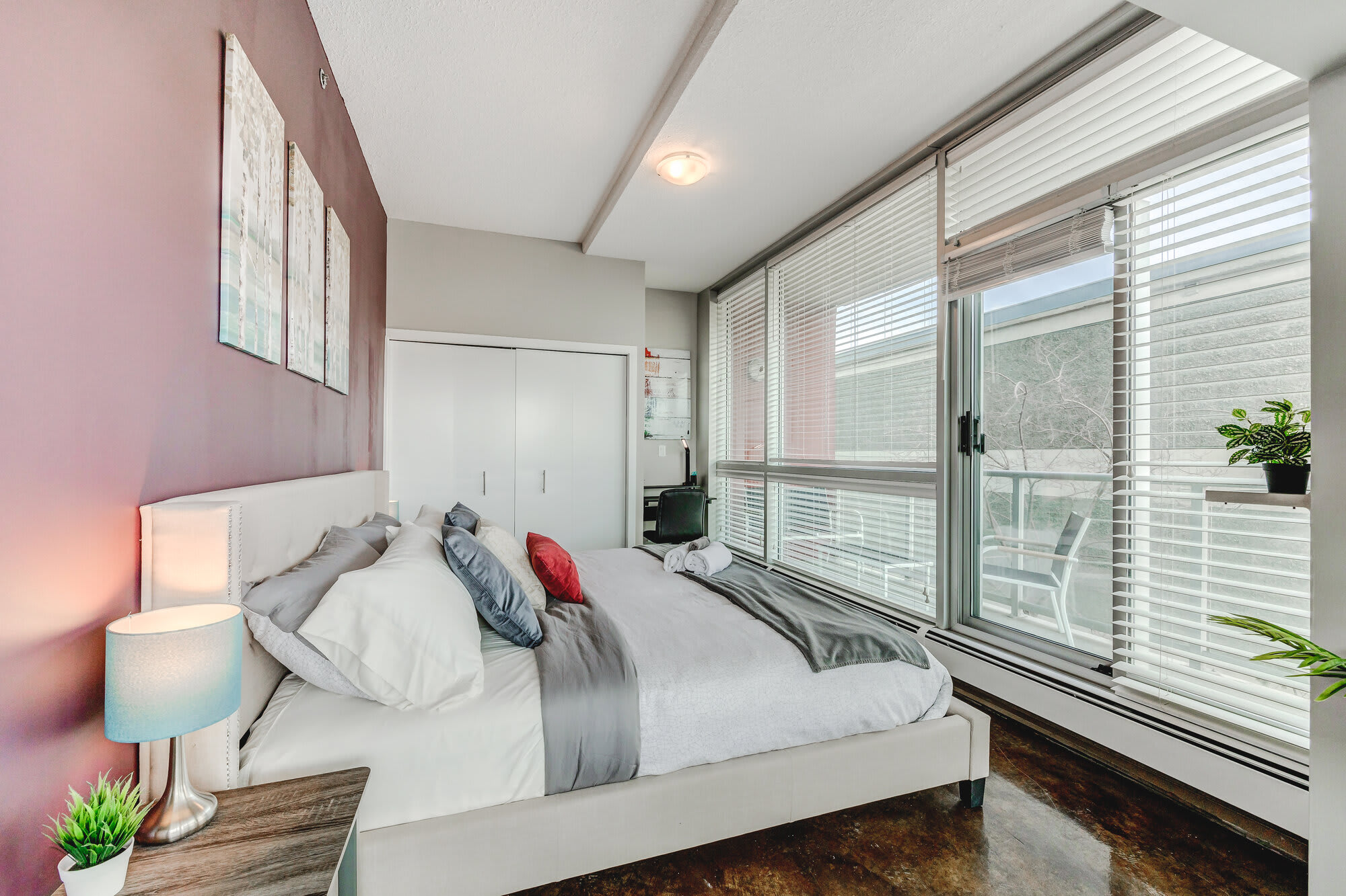 Spacious bedroom with a white upholstered bed, a nightstand, and a desk, featuring a plum accent wall. Large windows and a glass door provide access to a private balcony.