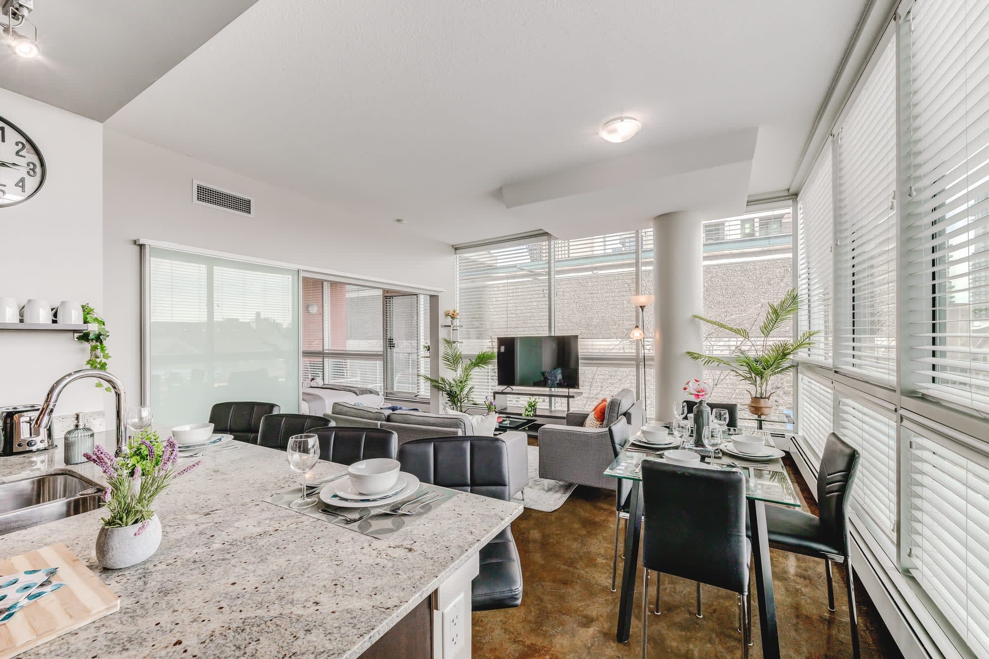 Open-concept living space showcasing a dining area with a glass table, a comfortable living room with a gray sofa and television, and a partial view of the kitchen island. Expansive windows with blinds provide ample natural light.