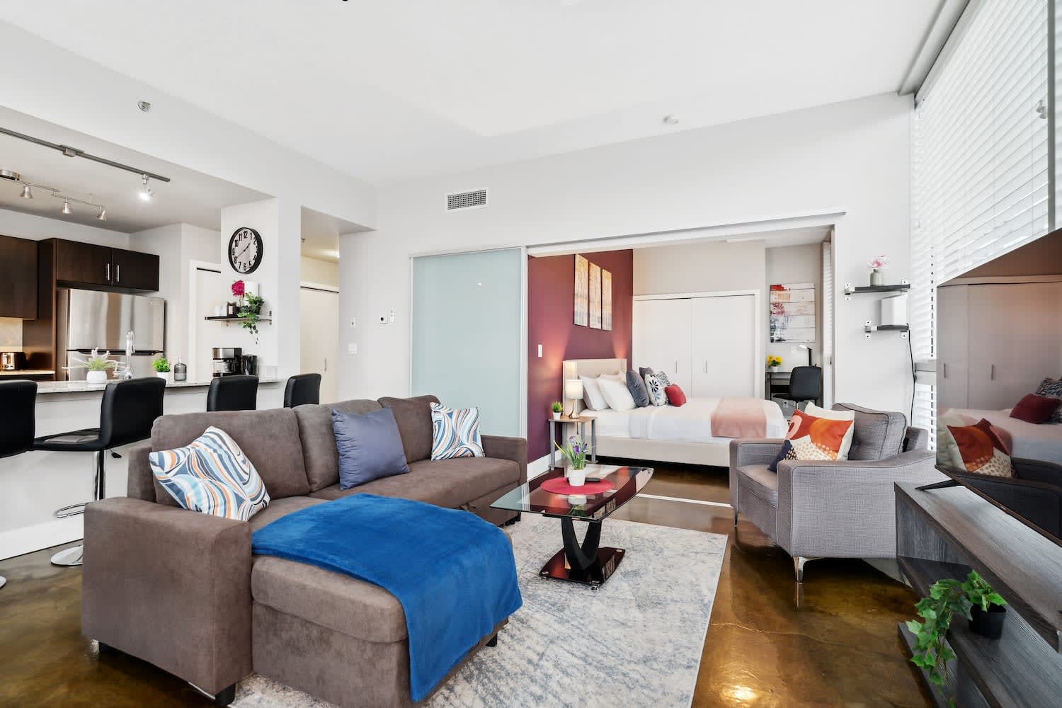 Open-concept living room with a brown sectional sofa and a glass coffee table, connected to a bedroom with a plum accent wall via a sliding frosted glass door. A kitchen is visible in the background.