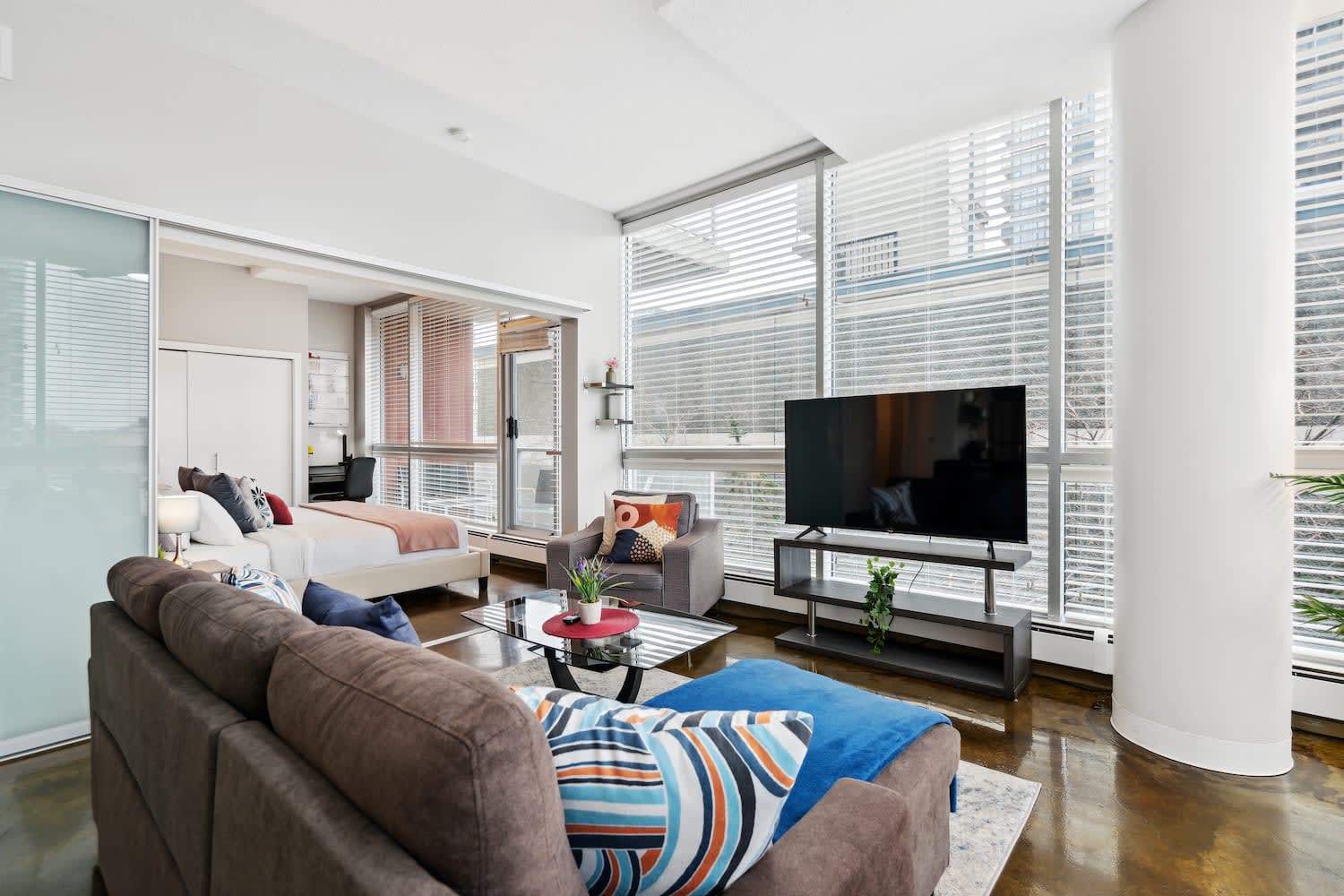 Modern living room with a brown sectional sofa and a grey armchair, facing a large flat-screen TV. A bedroom with a plum accent wall is visible through a wide opening, featuring a white bed.