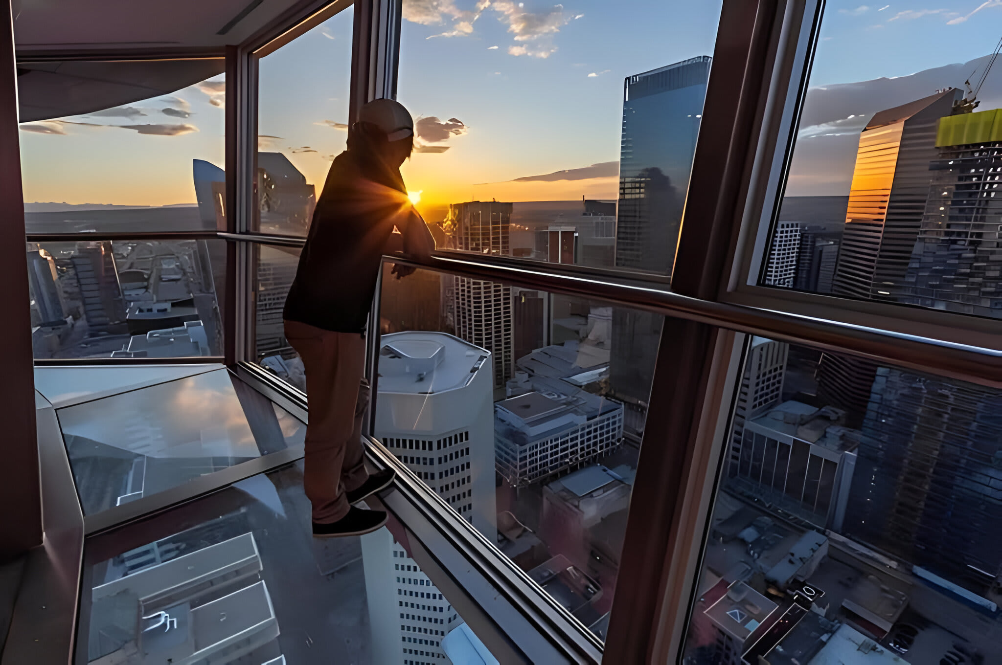 A person stands on a glass observation deck, gazing at a panoramic city skyline bathed in the warm glow of sunset, with numerous high-rise buildings below.