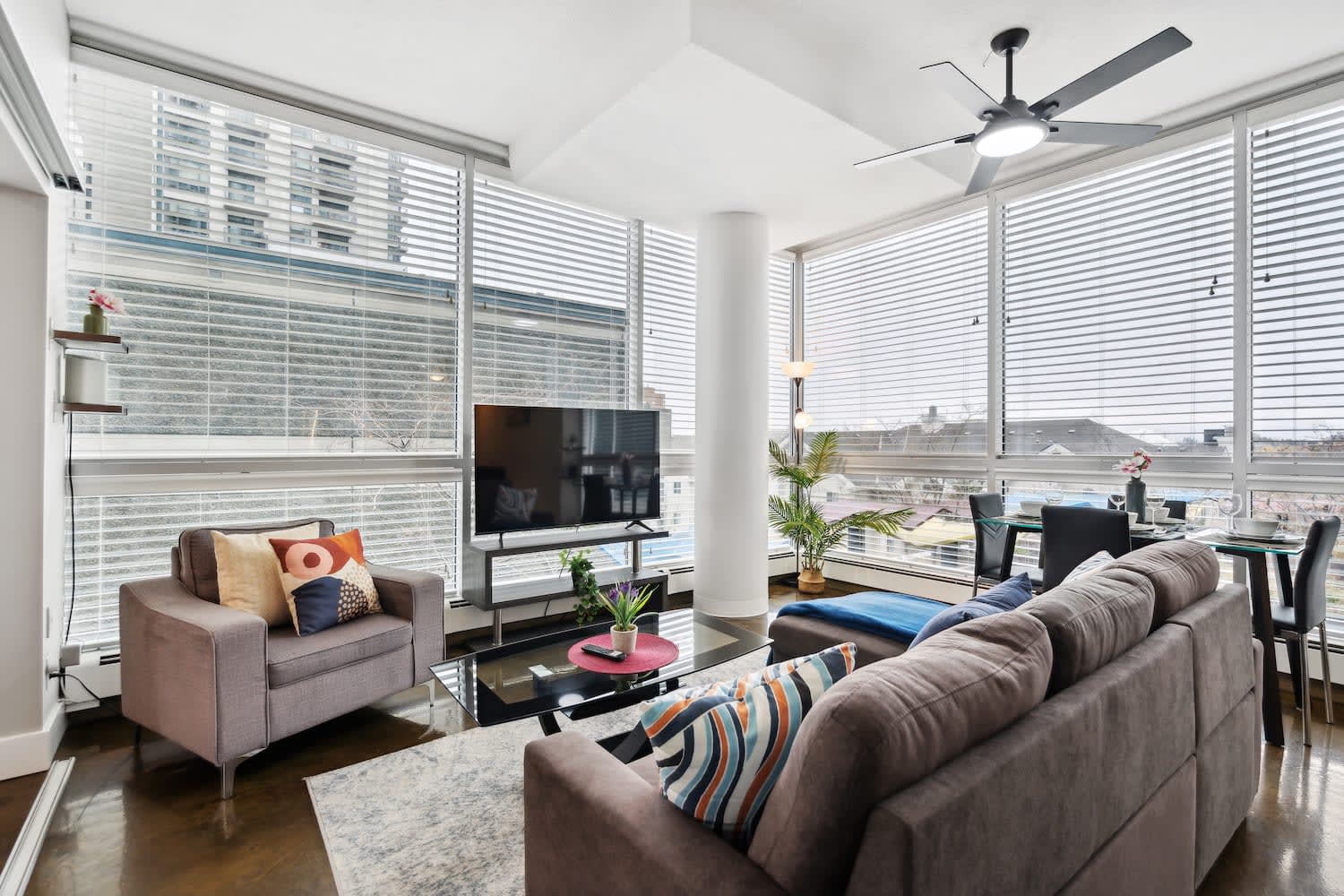 Bright living room with a grey armchair and a large flat-screen TV, featuring floor-to-ceiling windows with horizontal blinds that provide ample natural light and city views. A dining area is visible to the right.