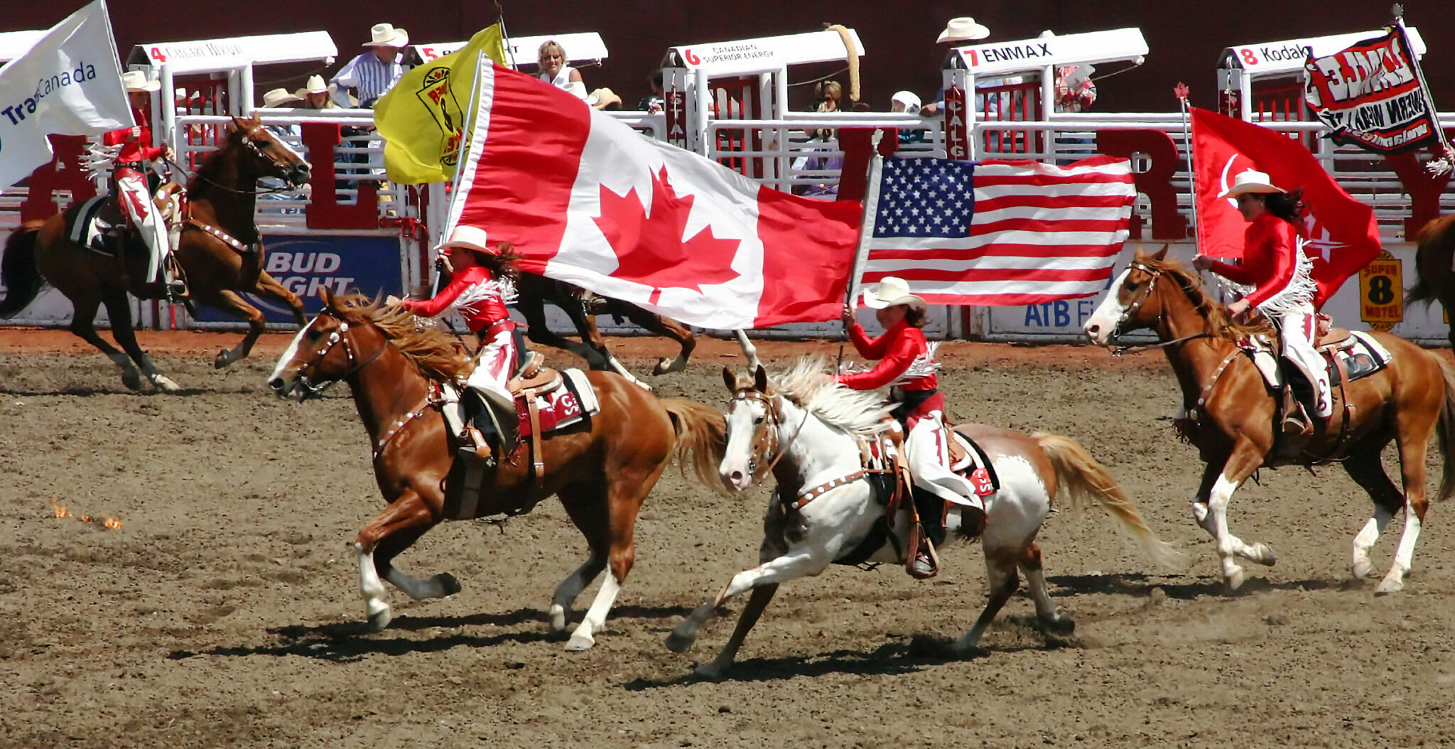 Rodeo riders on horseback gallop across a dirt arena, carrying large Canadian and American flags, with spectators visible in the stands in the background.