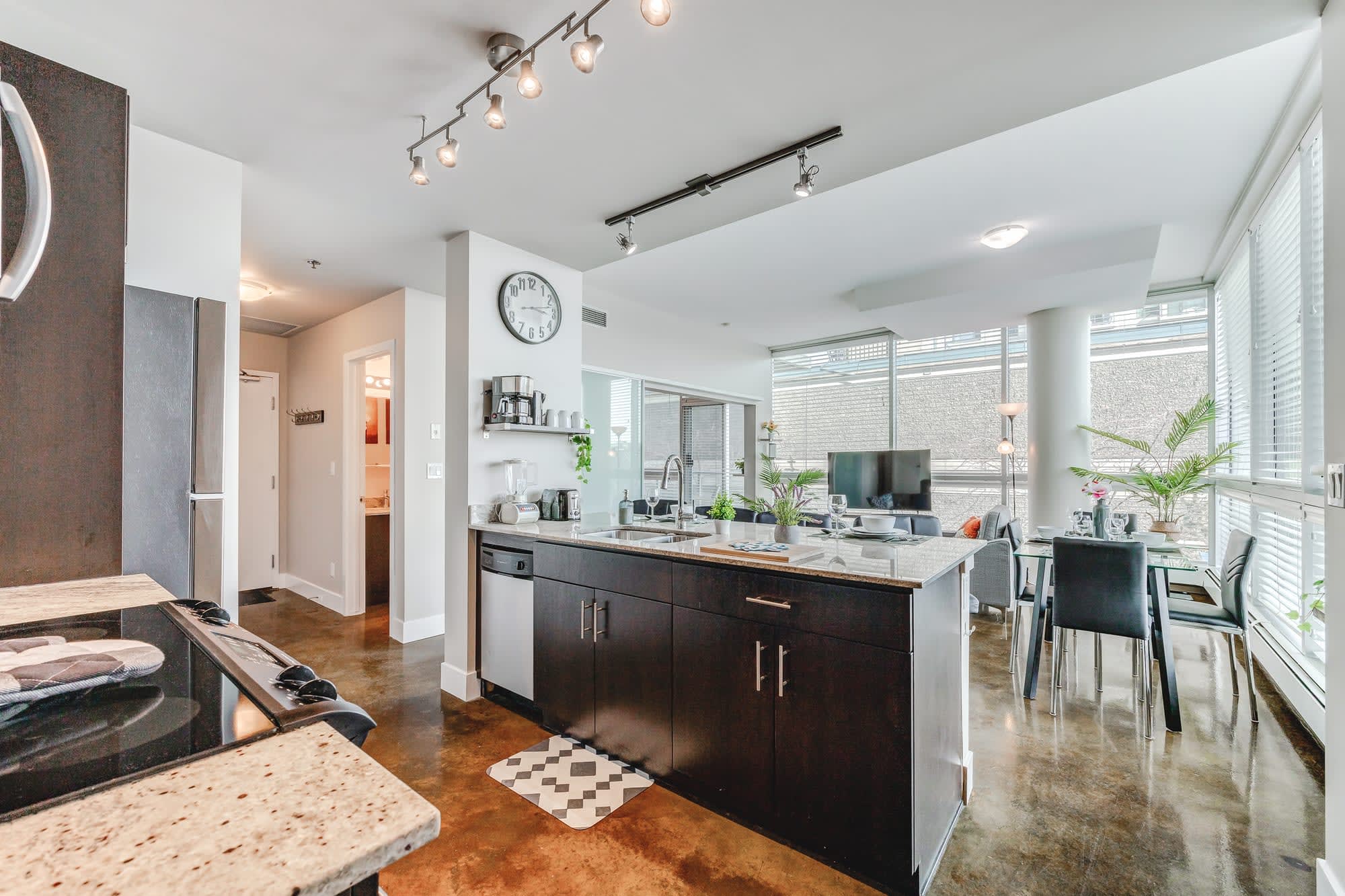 Sleek kitchen with dark wood cabinetry, granite countertops, and stainless steel appliances. A large island with a sink and seating defines the space, illuminated by modern track lighting.