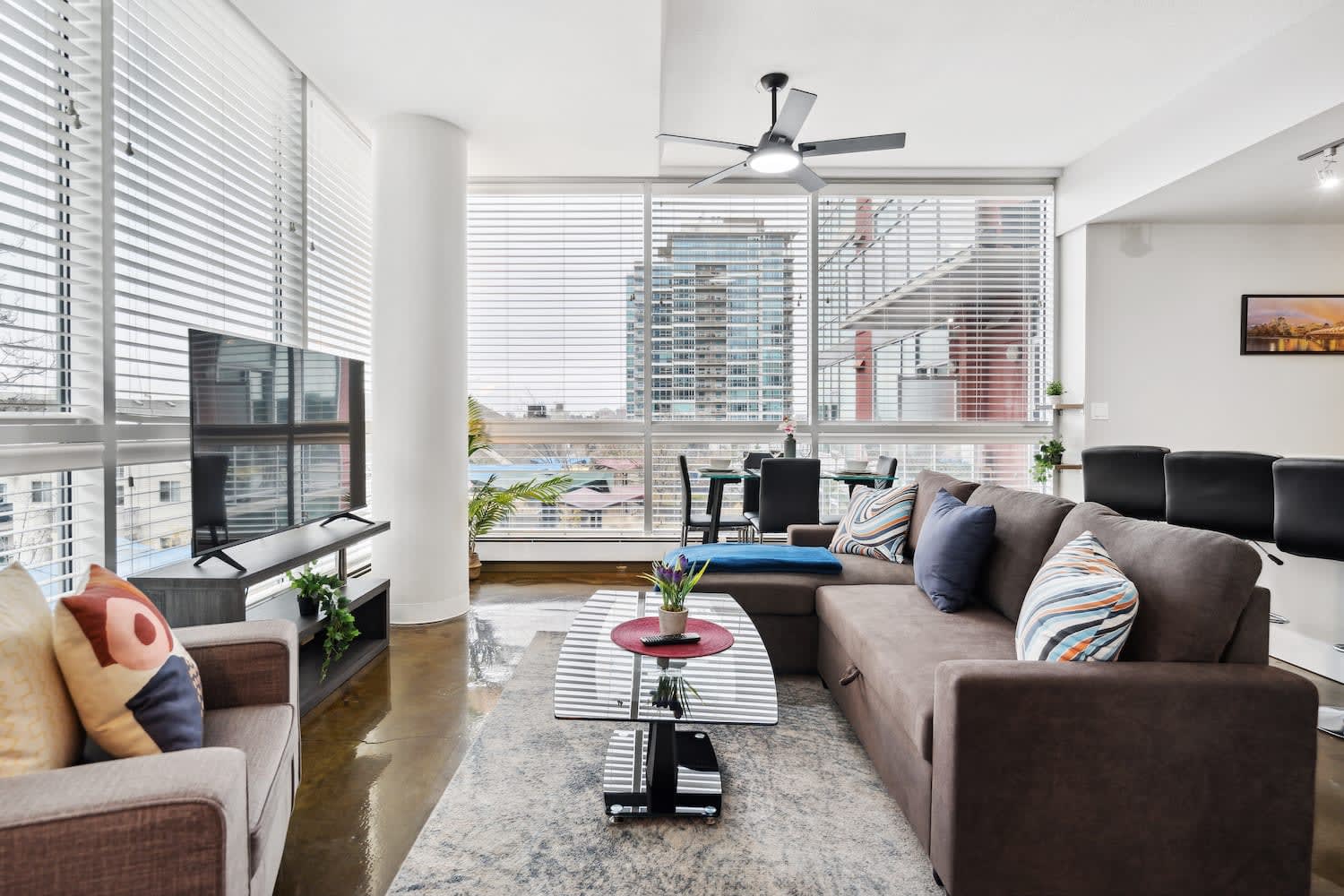 Contemporary living room with a brown sectional sofa, a grey armchair, and a large flat-screen TV on a media console. Expansive windows with blinds offer city views, and a ceiling fan is overhead.