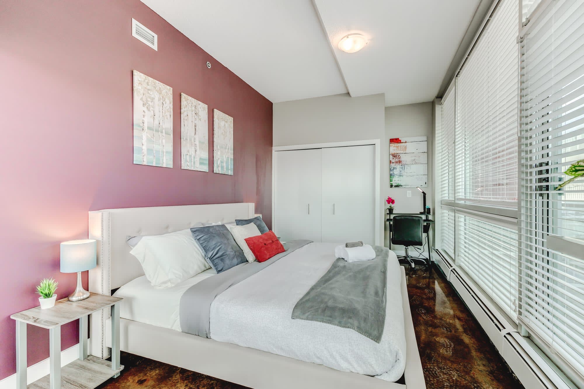 Bright bedroom featuring a white upholstered bed, a rustic nightstand with a lamp, and a small desk area. The room boasts a plum accent wall, white sliding closet doors, and large windows with white blinds.