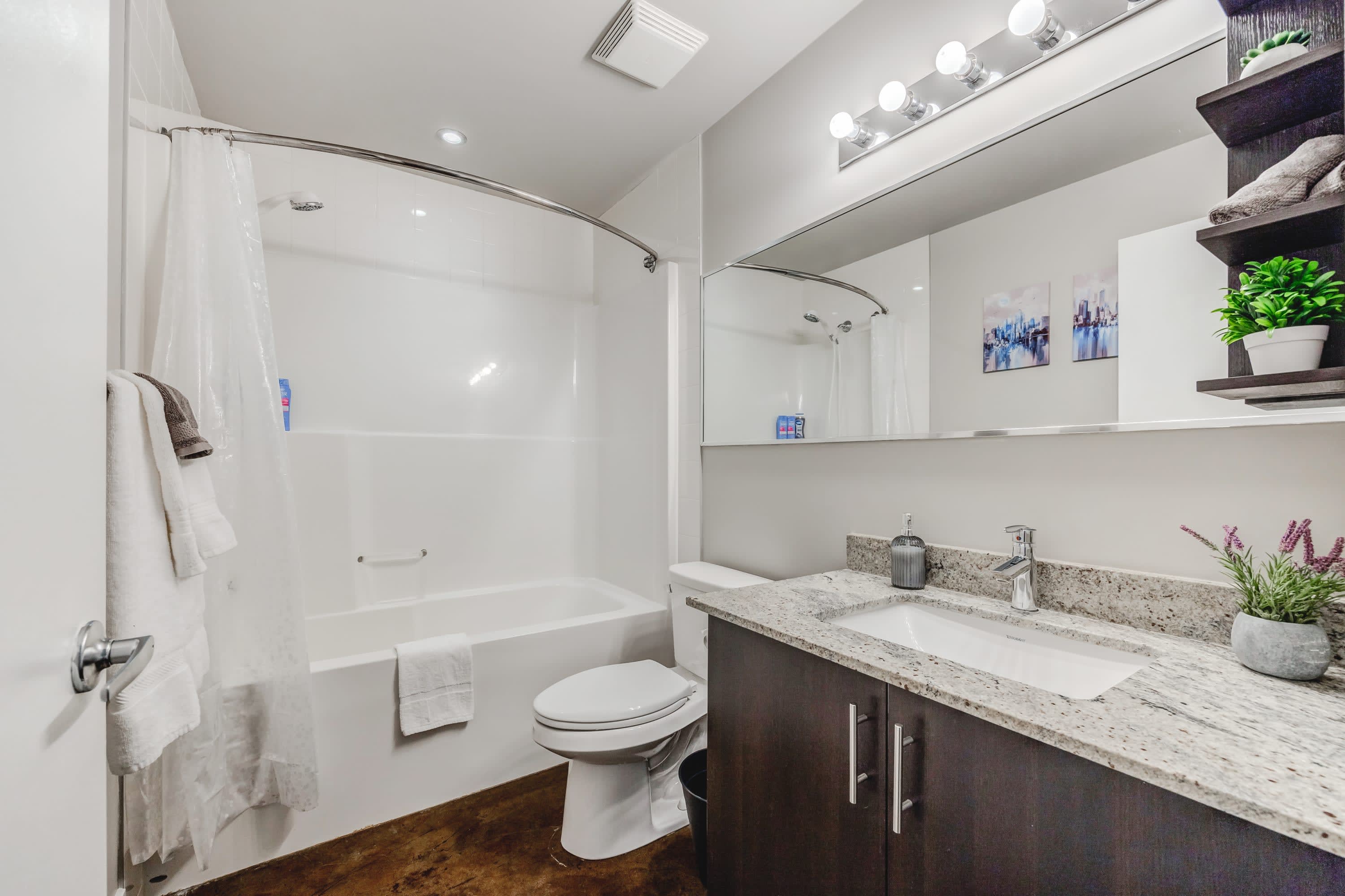 Full bathroom equipped with a white shower-over-tub combo, a toilet, and a single vanity with a granite countertop and dark wood cabinet. A large mirror and modern light fixture are present.