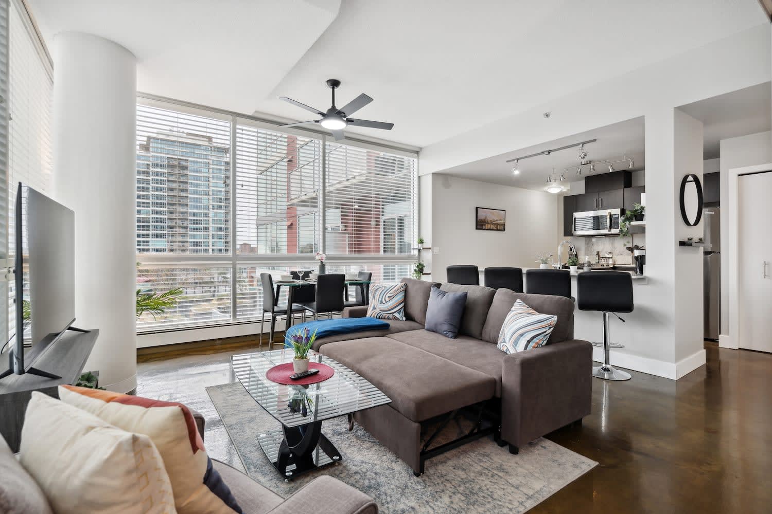 Modern living room featuring a brown sectional sofa with a pull-out bed mechanism visible, a glass coffee table, and large windows with blinds overlooking city buildings. A dining area and kitchen are in the background.