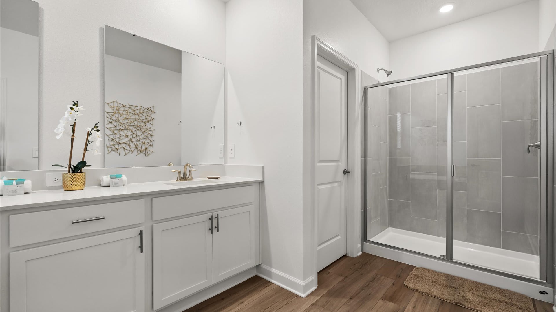 Modern bathroom featuring a white single vanity with a large mirror, gold-toned faucet, and a glass-enclosed walk-in shower with light gray subway tiles. The floor is light wood-look planking.