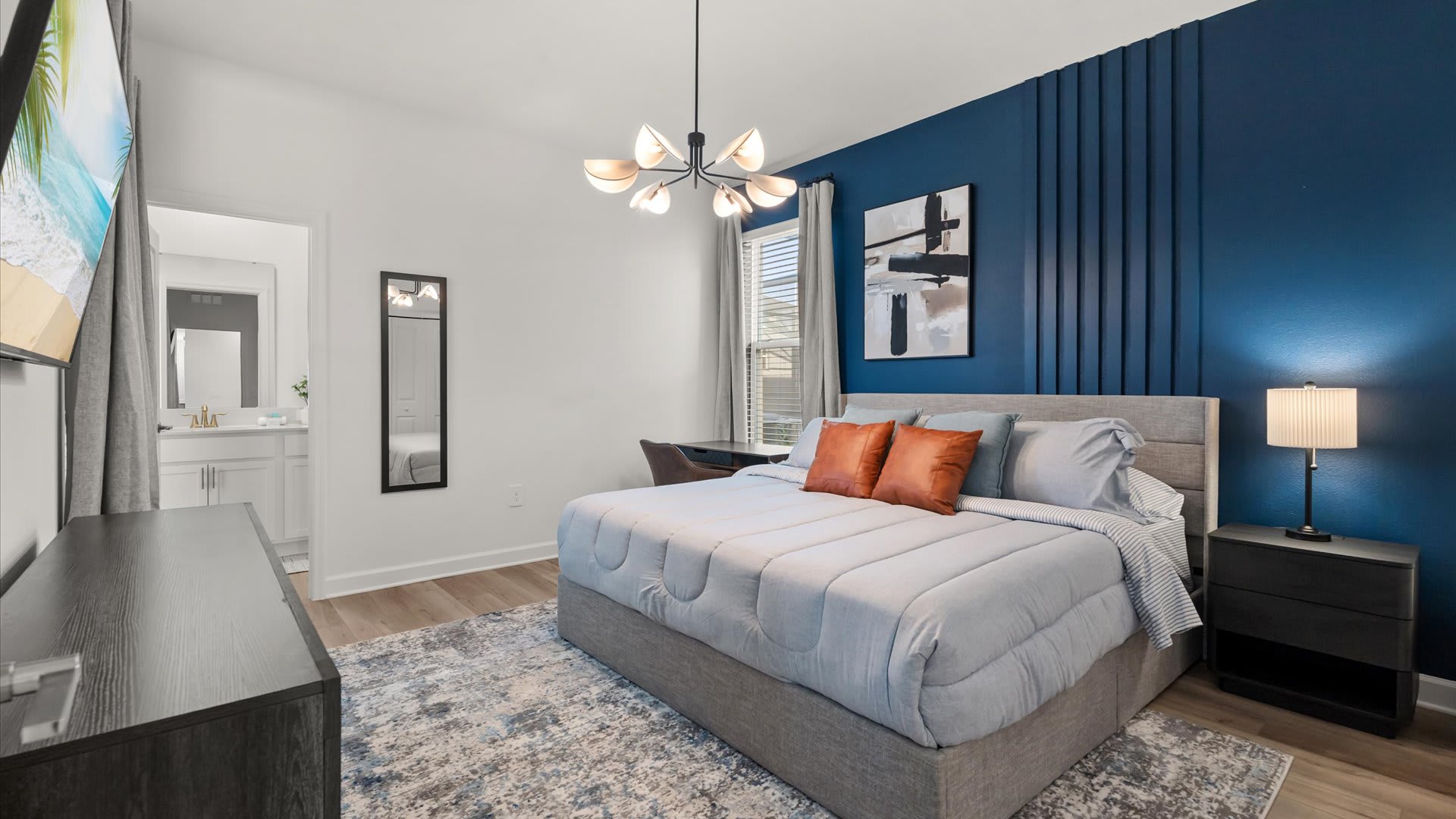 Stylish bedroom with a queen-sized bed, a striking dark blue accent wall with vertical paneling, and a modern chandelier. A desk and chair are visible by the window, and a flat-screen TV is mounted on the opposite wall.