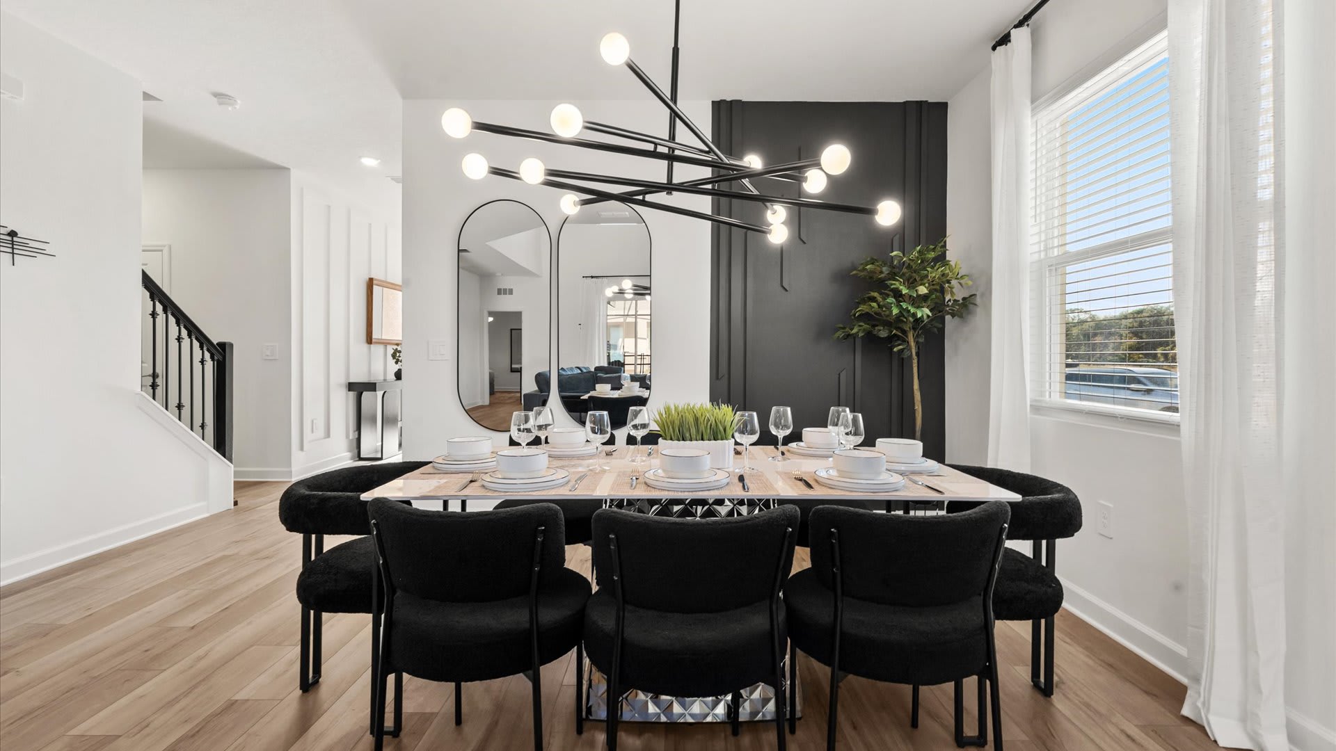 Close-up of the modern dining area featuring a long wooden table set for eight with black chairs, illuminated by a unique linear chandelier, with a dark accent wall and decorative mirrors in the background.
