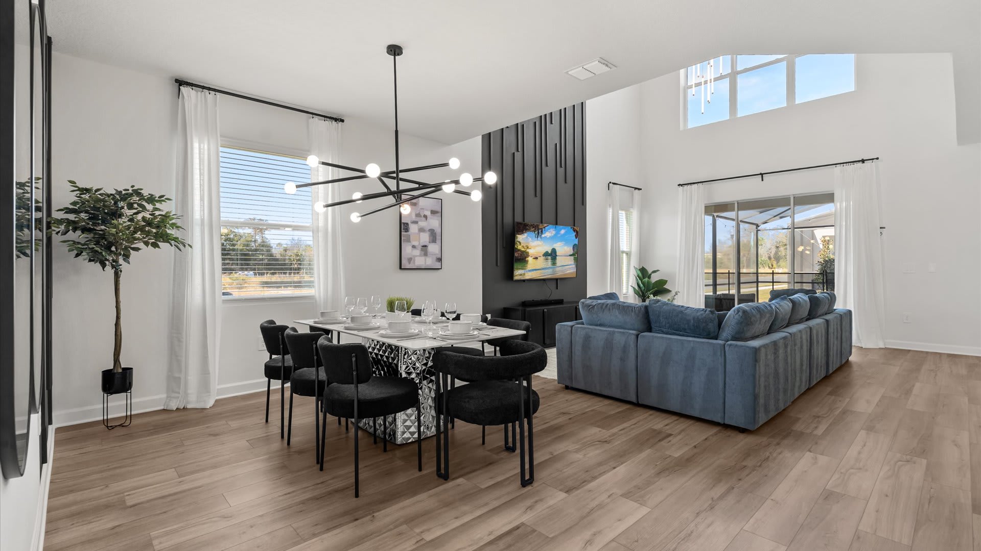 Open-concept dining area with a long rectangular table set for eight, black chairs, and a striking modern chandelier, adjacent to a living room with a blue sectional and a TV mounted on a dark accent wall.