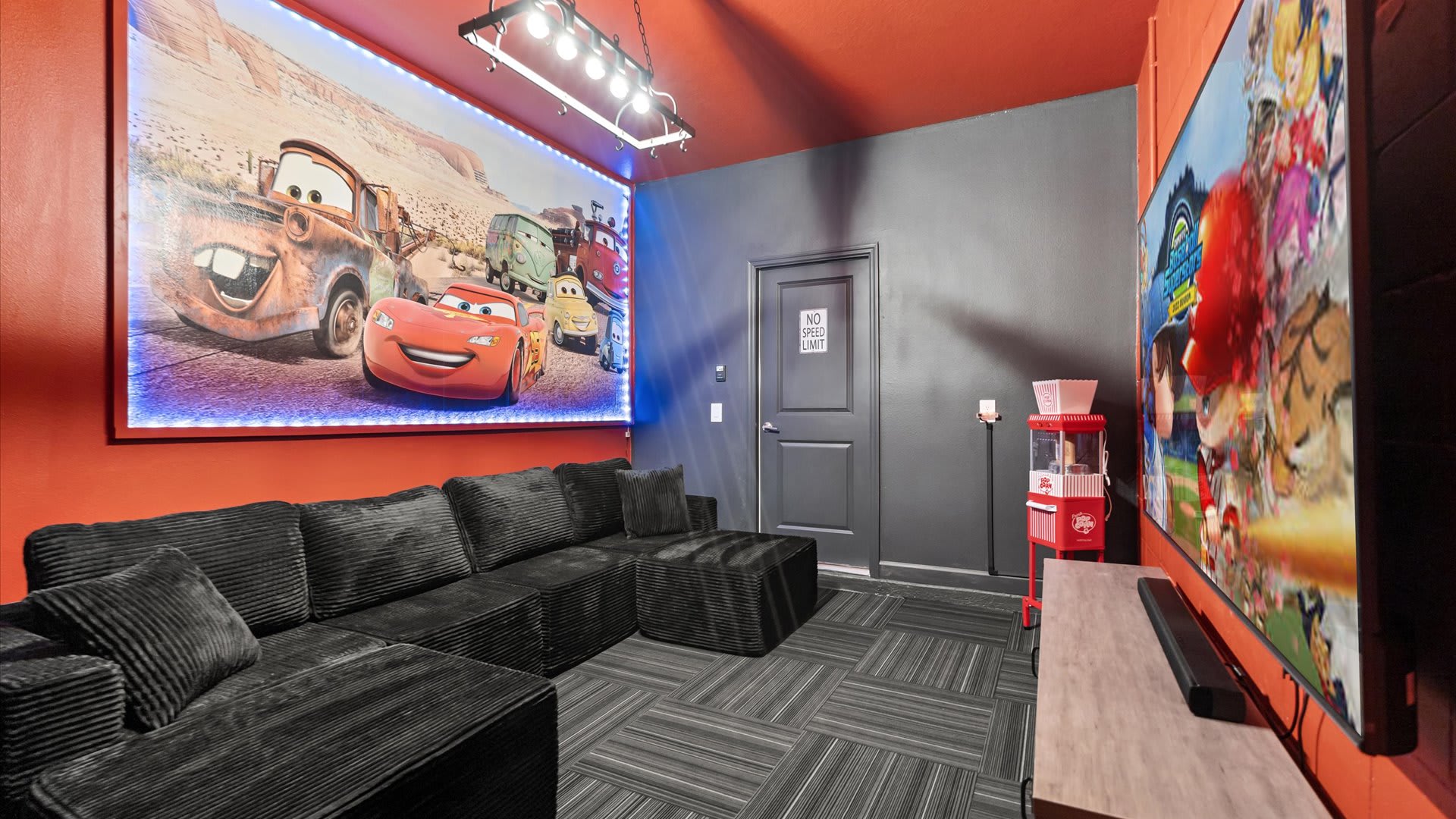A vibrant media room features a large L-shaped black sectional sofa facing a big backlit mural of 'Cars' characters, with a popcorn machine to the right. The walls and ceiling are painted a bold orange, contrasting with the dark gray door and patterned carpet.