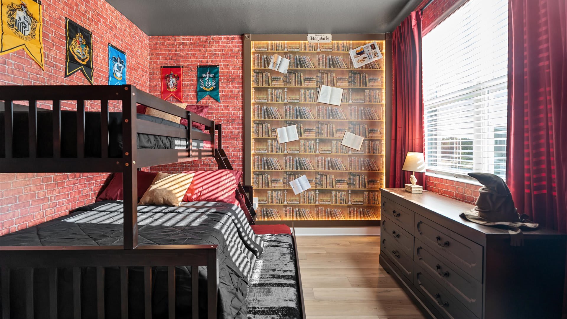Another view of the Harry Potter themed bedroom, showcasing the dark wood bunk beds and the illuminated bookshelf mural on the wall. Harry Potter house banners and a sorting hat on the dresser add to the theme.