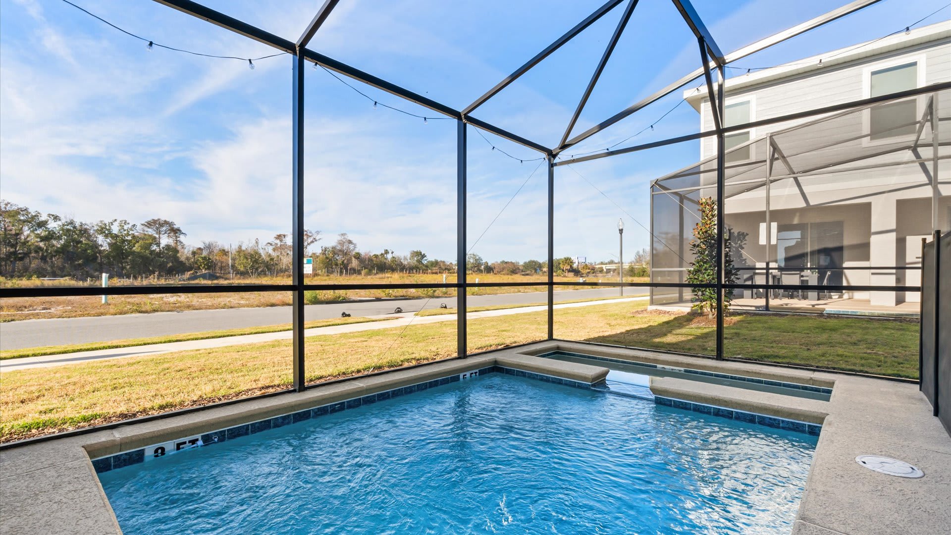 A wide shot of the screened-in private pool area shows a clear blue swimming pool and hot tub, with string lights overhead. The enclosure offers views of a grassy area and a road, with another house visible in the distance.