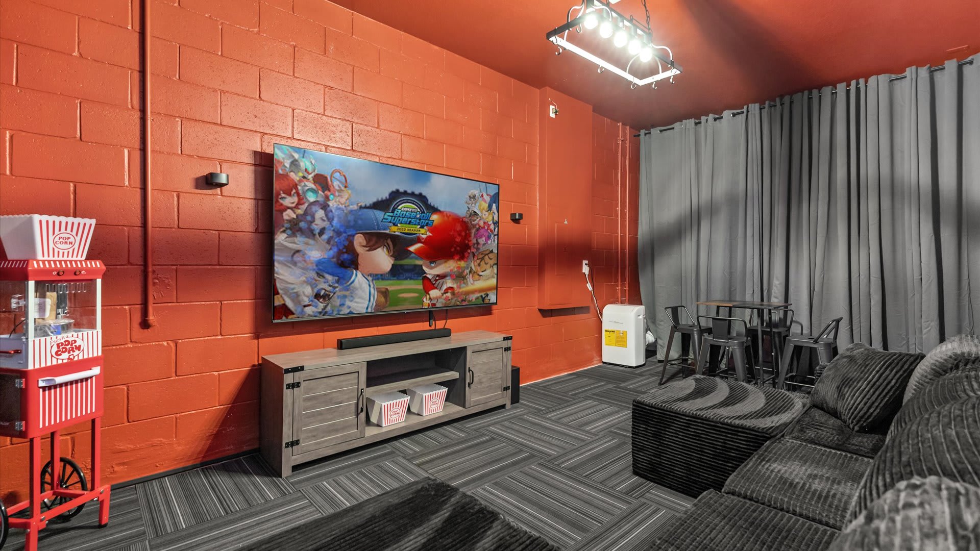 A vibrant media room with orange brick walls features a large flat-screen TV mounted above a wooden console, with a popcorn machine to the left. A dark gray sectional sofa provides seating, and a small high-top table with stools is visible in the corner.