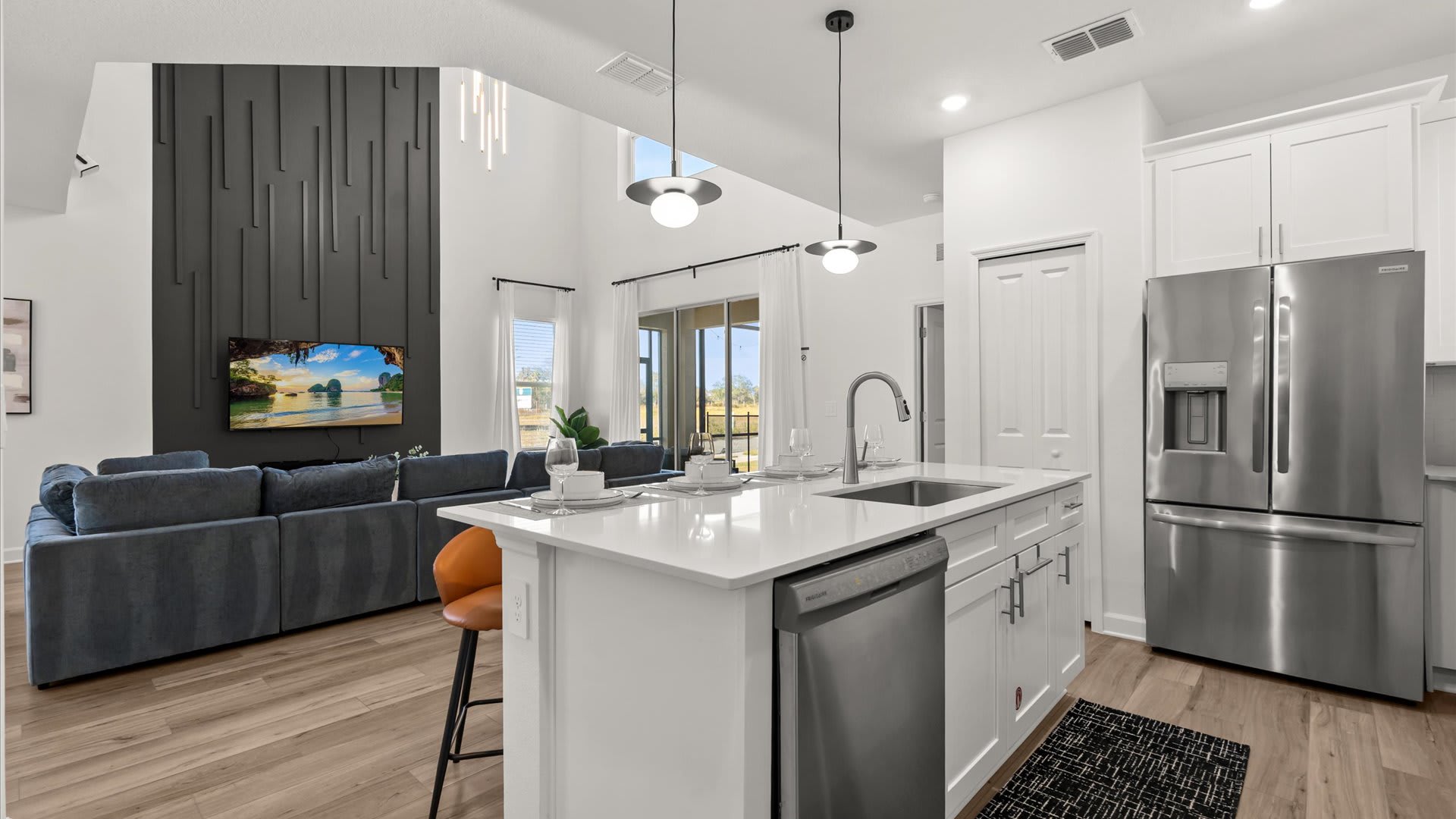 View from the kitchen island looking into the open-concept living area, showing white shaker cabinets, stainless steel appliances, and a large blue sectional sofa in the background with a TV on a dark accent wall.