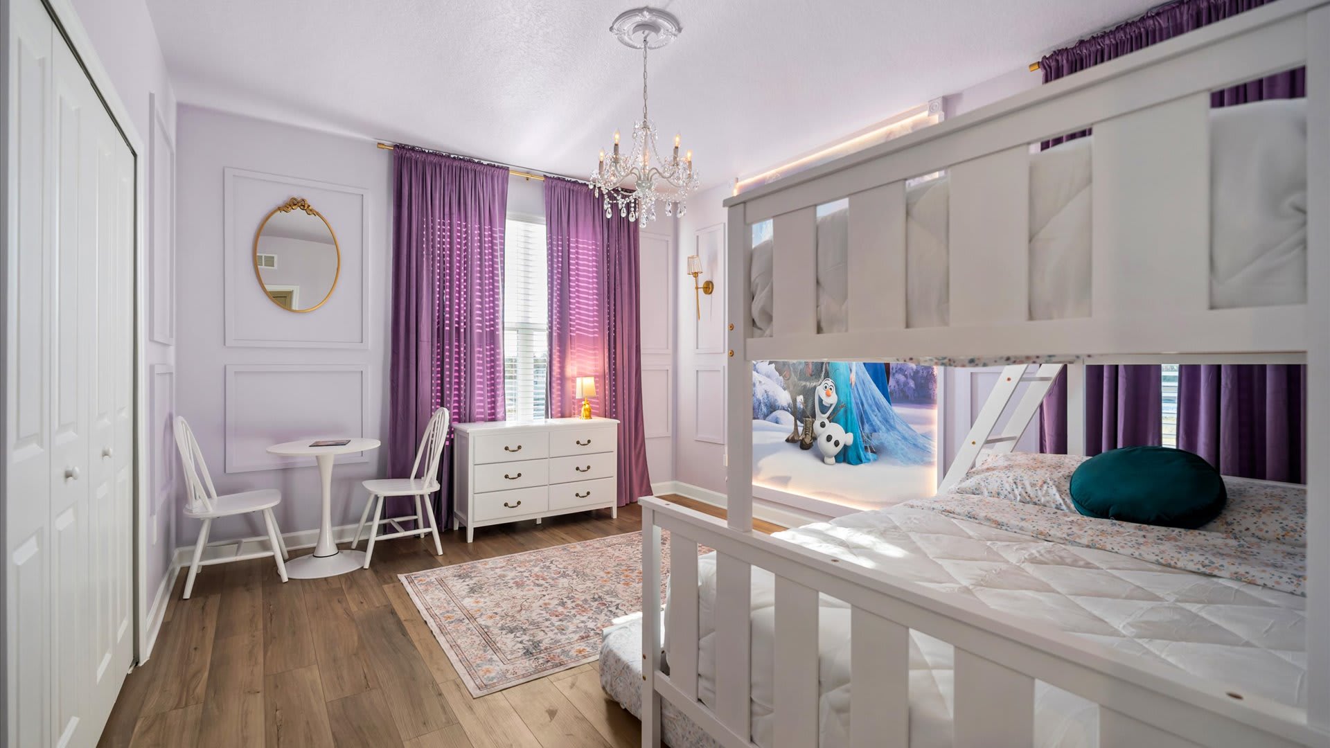 Another view of the kids' bedroom with white bunk beds, a crystal chandelier, and light purple walls. A white dresser, a small table with chairs, and a gold-framed mirror are visible.