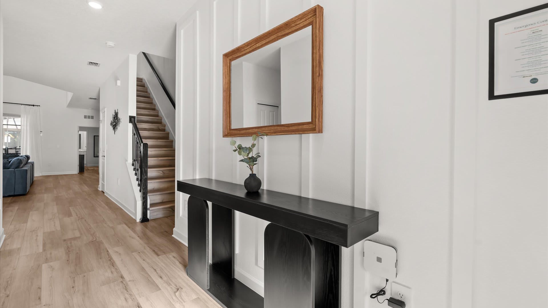 Bright entryway hallway with light wood-look flooring, a black console table with a vase and mirror, and white wainscoting on the wall, leading towards a staircase with black railings.