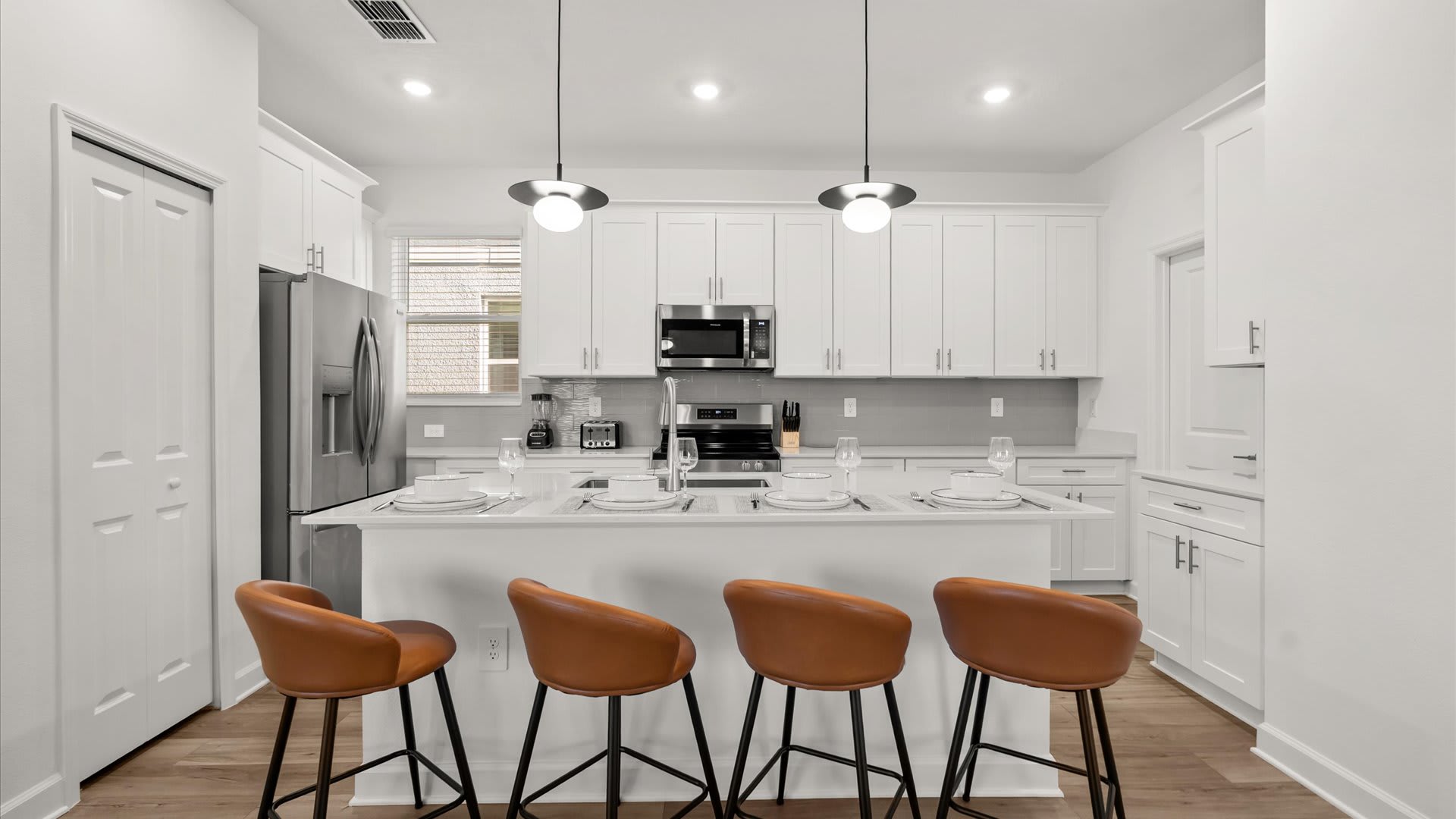 Contemporary kitchen island with four brown leather bar stools, a sink, and pendant lighting, surrounded by white shaker cabinets and stainless steel appliances, with a window providing natural light.
