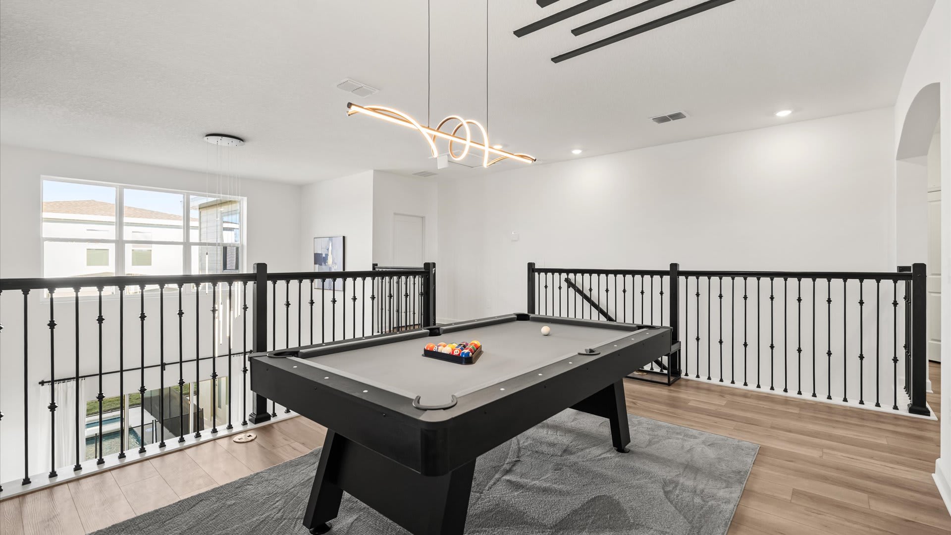 A bright game room in a loft area features a gray-felted pool table with cues, positioned on a gray area rug. The space is open to the lower level, with black metal railings and a modern linear chandelier overhead.