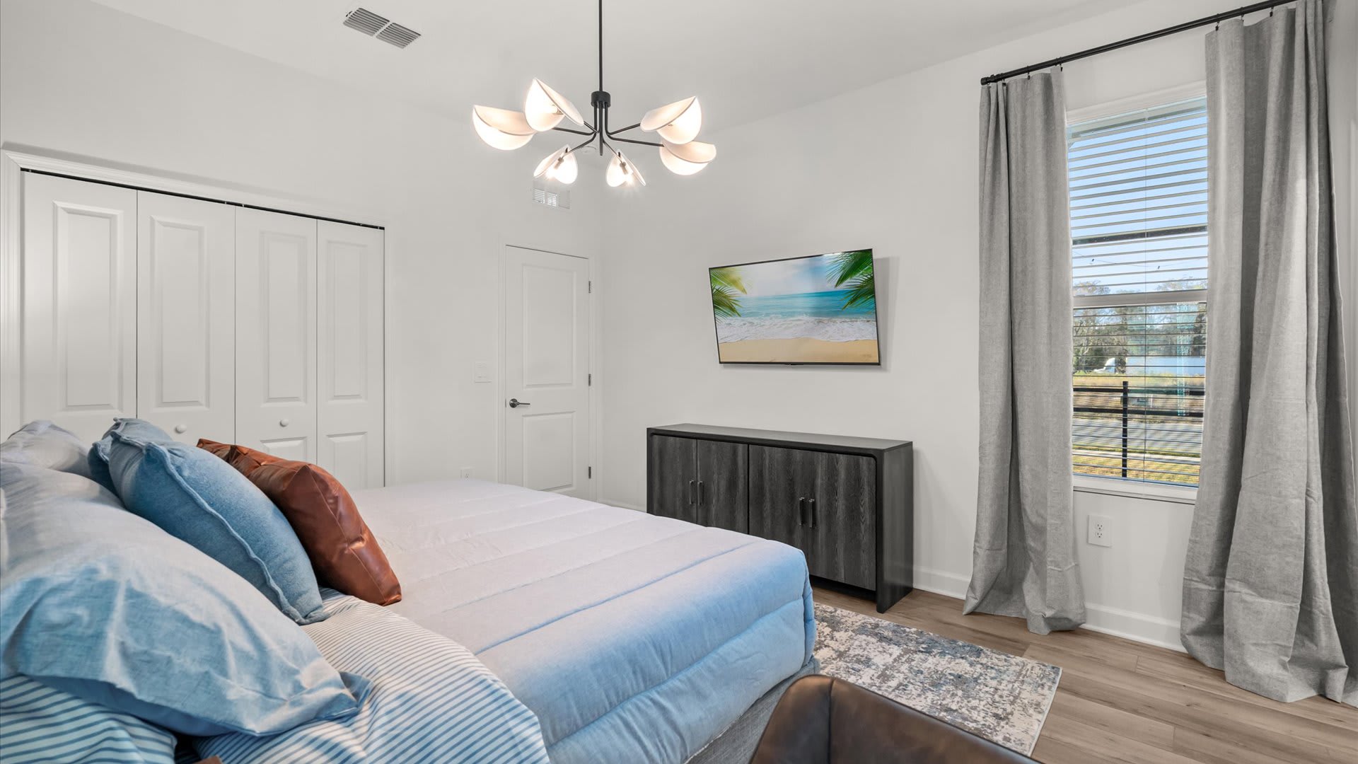 View of the bedroom from the foot of the bed, showing a wall-mounted flat-screen TV above a dark wood console. A large window with blinds and gray curtains offers a view of the outdoors.