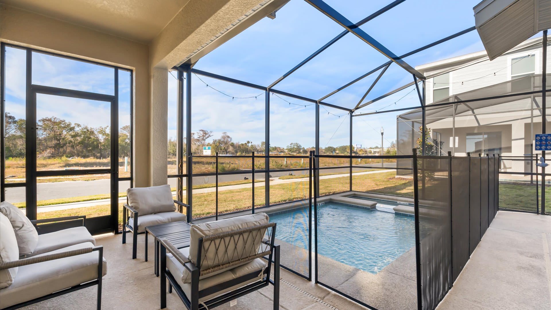 A covered outdoor patio area, enclosed by a screen, features comfortable outdoor seating with light-colored cushions. The patio overlooks a private swimming pool and hot tub, which are separated by a safety fence.