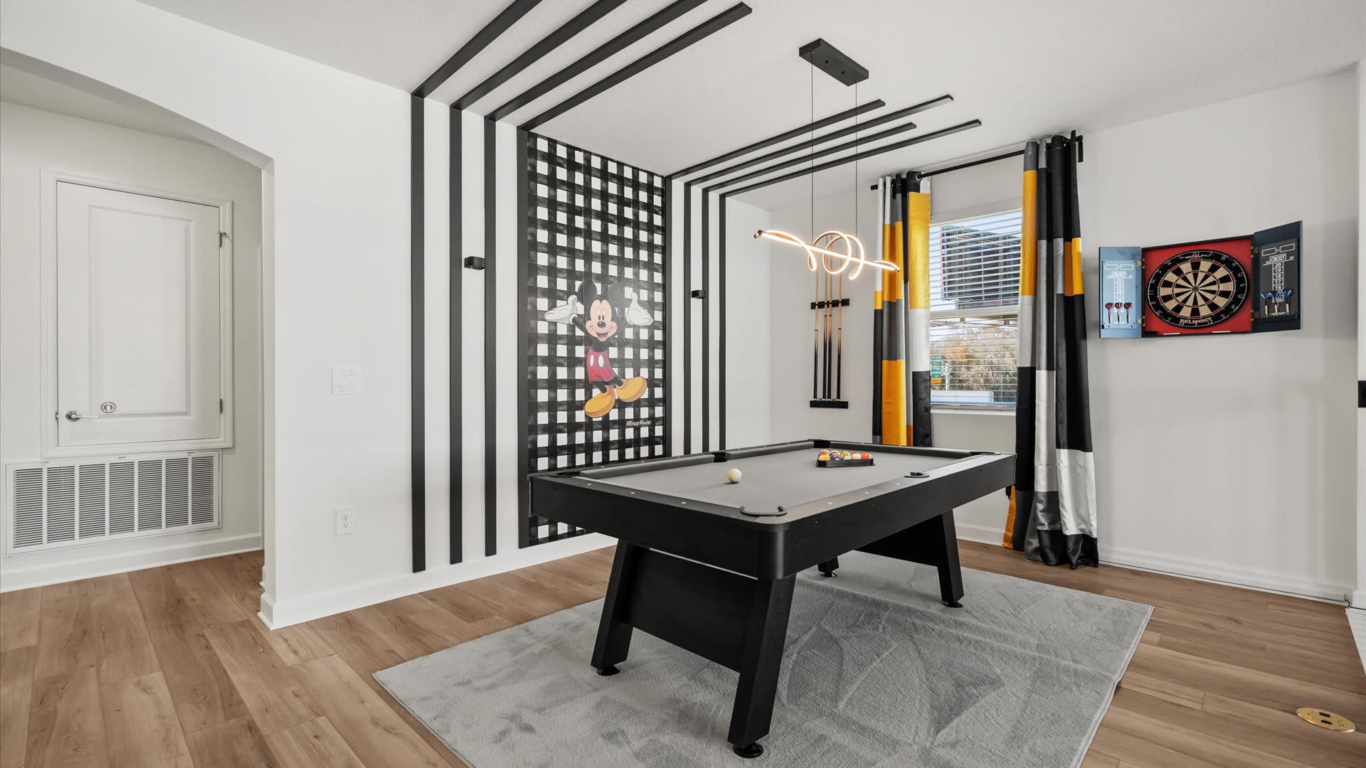 A game room features a gray-felted pool table with cues, set against a white wall with a black and white striped accent and a Mickey Mouse graphic. A dartboard is mounted on the adjacent wall, and a modern light fixture hangs overhead.