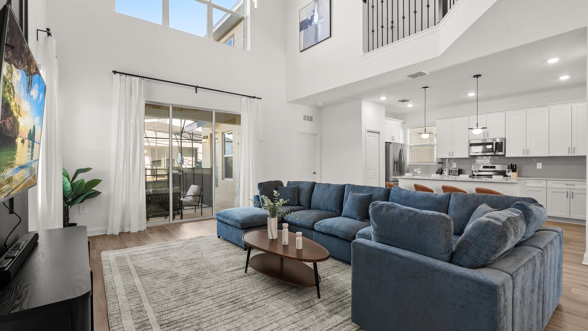 Bright, open-concept living room with a large blue sectional sofa, a modern coffee table, and a large TV mounted on the wall, featuring high ceilings and large sliding glass doors leading to an outdoor area.