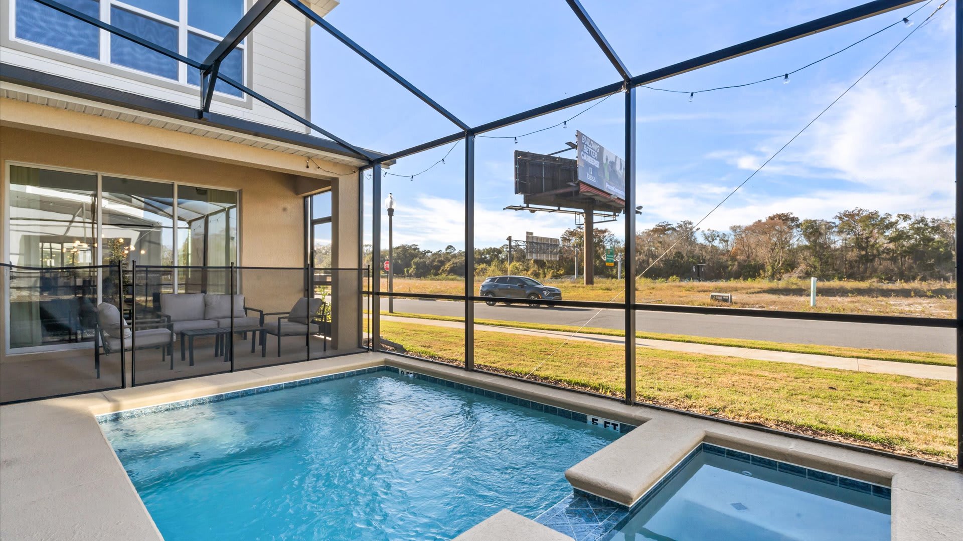 An outdoor screened-in pool area features a rectangular swimming pool with an integrated hot tub, surrounded by a safety fence. Beyond the screen, a road with a car and a billboard are visible, indicating a roadside location.