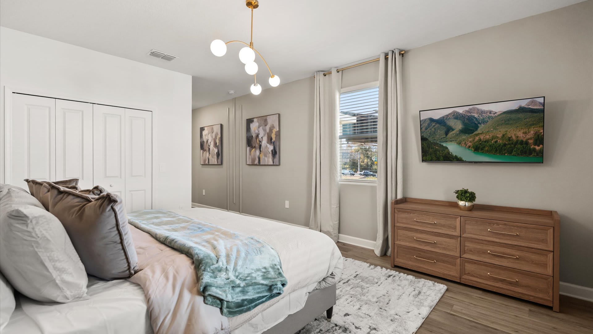 Cozy bedroom with a large bed featuring a grey headboard and blue throw, a wooden dresser with a TV mounted above it, and a modern gold chandelier, with a window providing natural light.