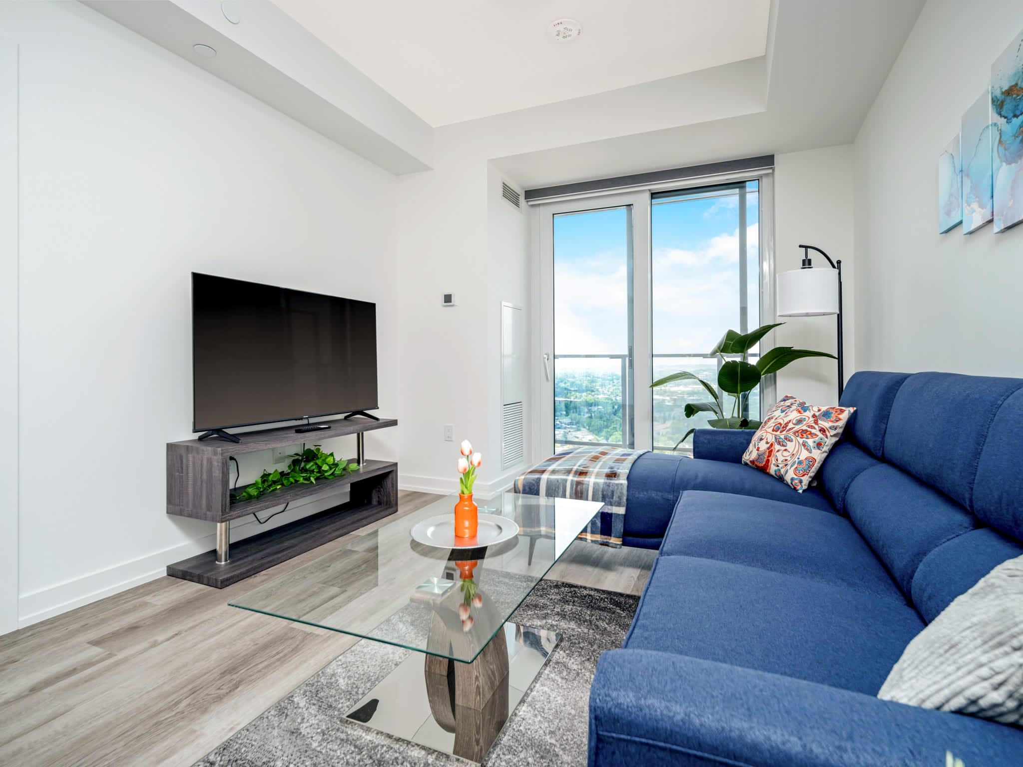 Cozy living room featuring a blue sectional sofa with decorative pillows, a glass coffee table, and a wall-mounted TV. A large sliding glass door provides access to a balcony with city views.
