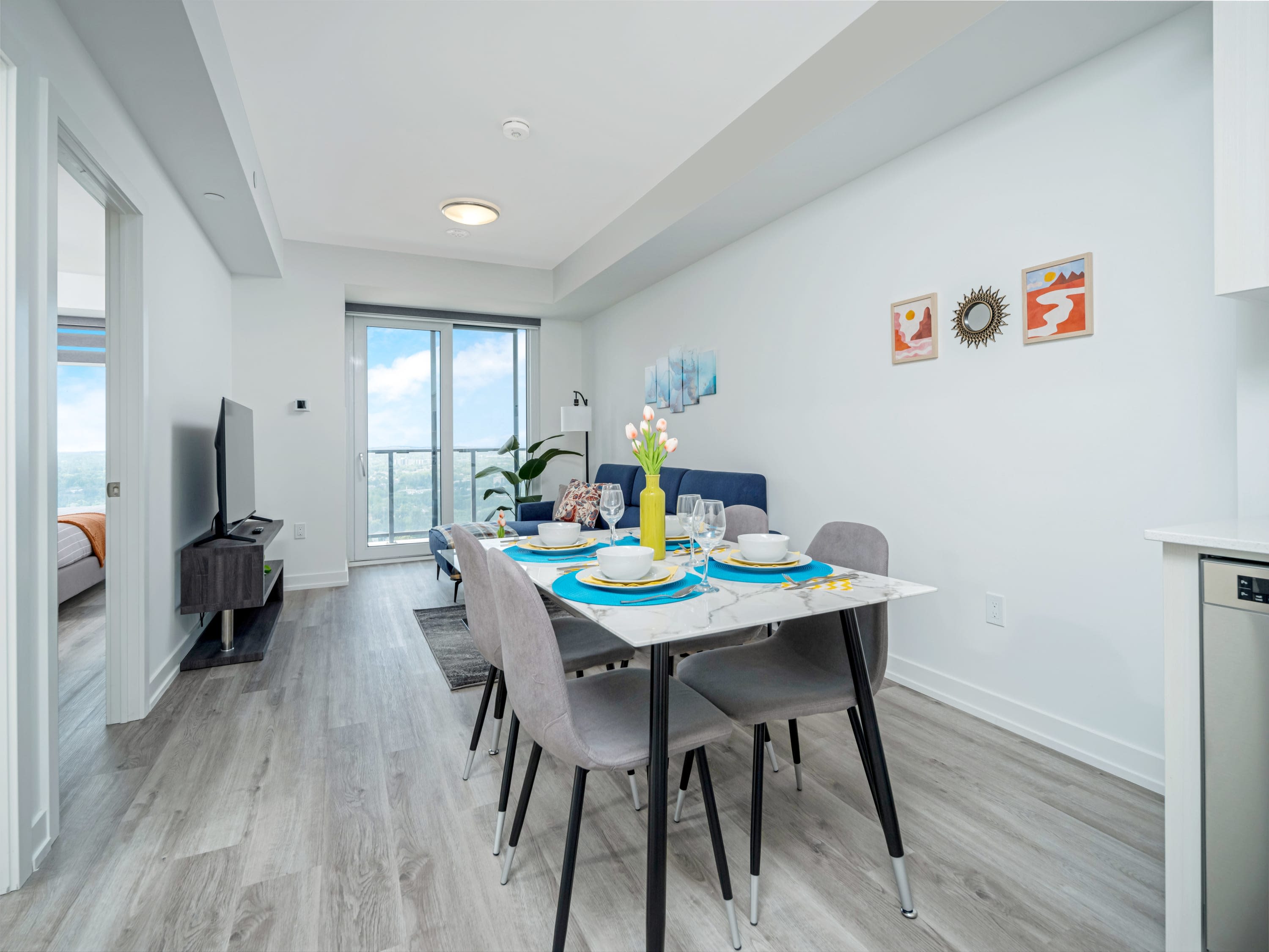 Open-concept living and dining area with light laminate flooring, featuring a blue sectional sofa, glass coffee table, and a dining table with four chairs. The space is brightly lit with natural light from the balcony door.