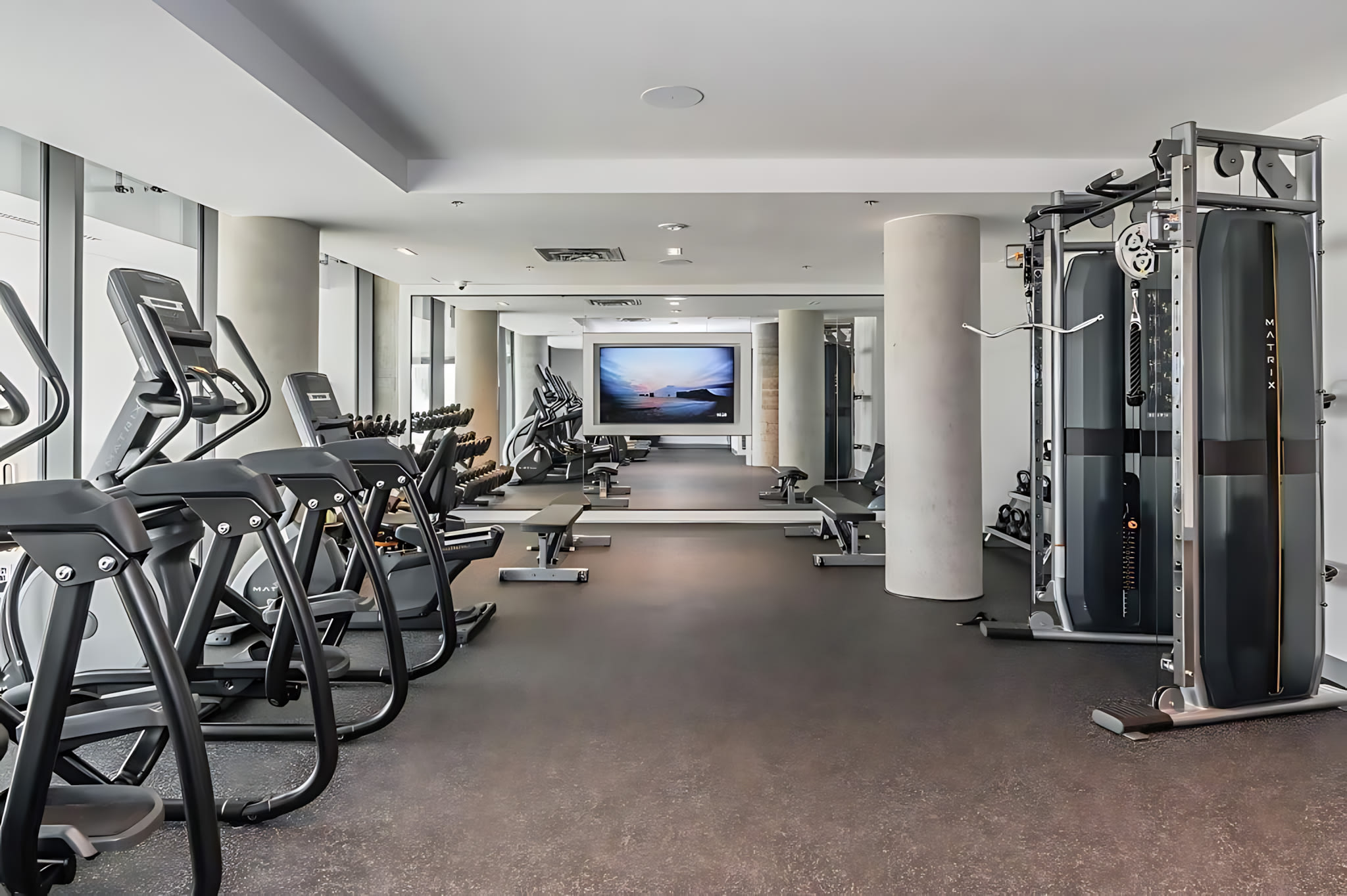 Well-equipped fitness center with various exercise machines including ellipticals, treadmills, and weight machines, along with free weights, and a large screen displaying a scenic view.