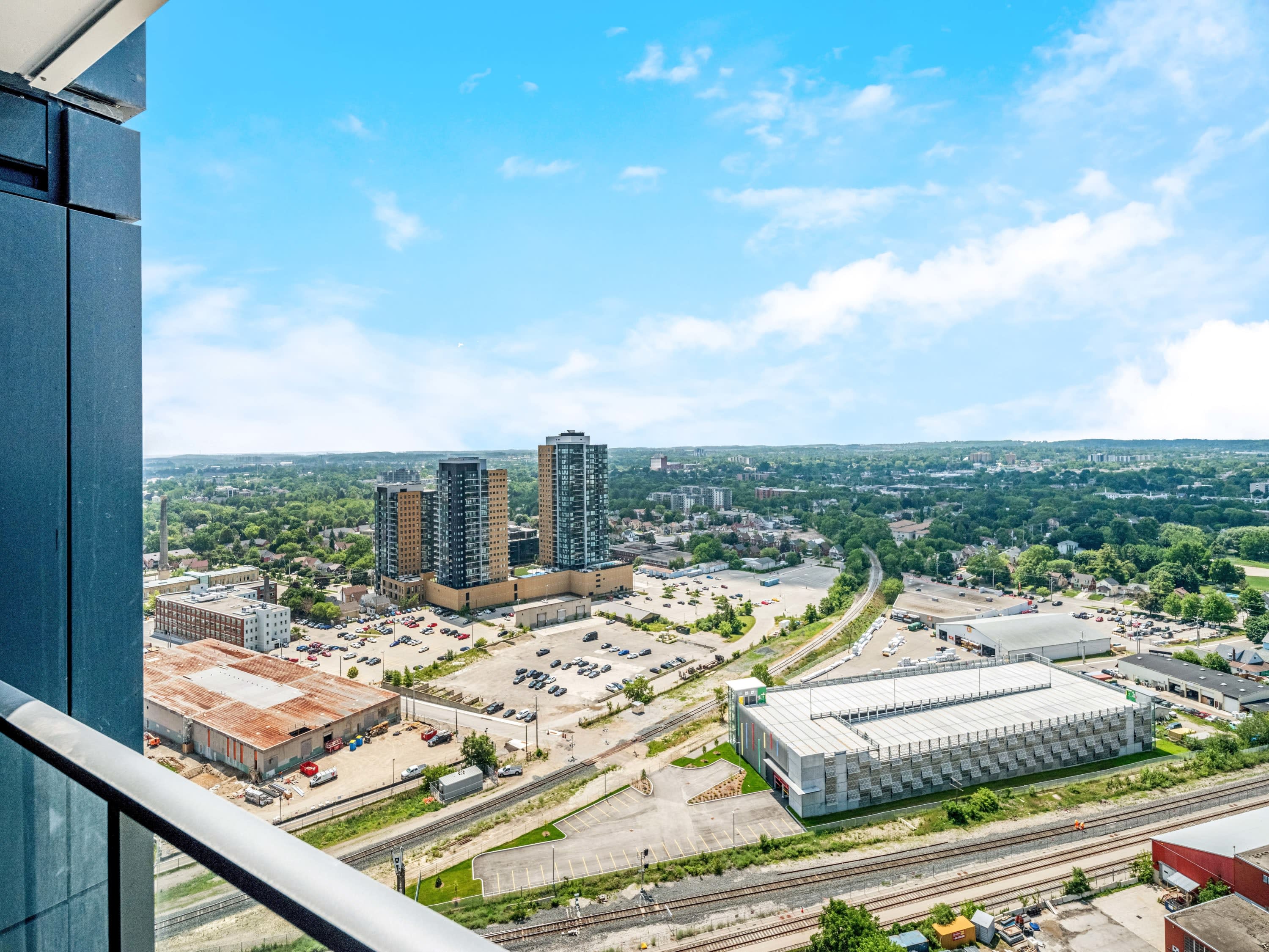 Expansive balcony view from a high floor, revealing a mix of urban and suburban development, including modern buildings, parking lots, and green spaces, all under a clear blue sky.