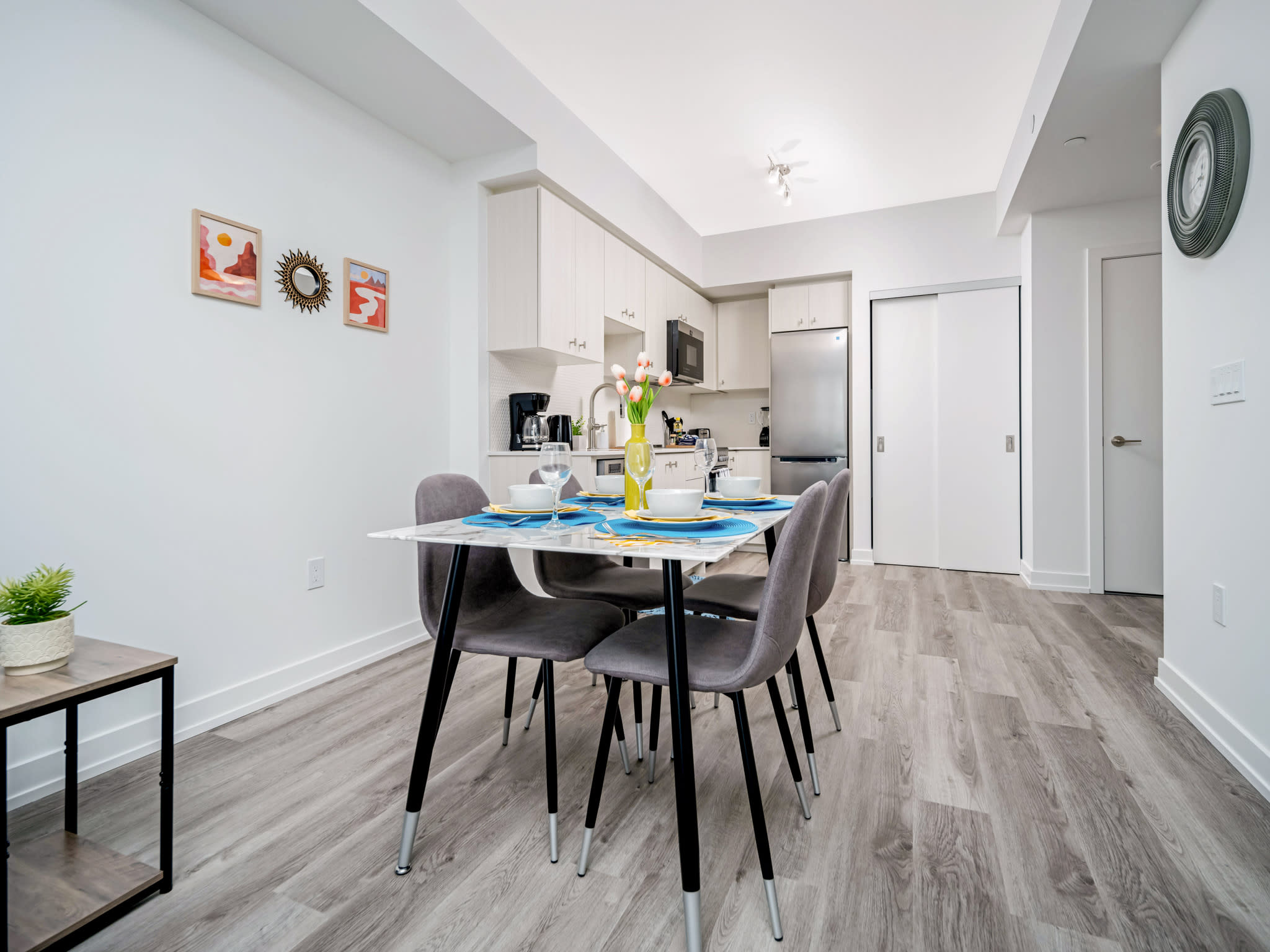 Modern dining area with a white rectangular table set for four, featuring gray upholstered chairs with black legs, light wood-look flooring, and a view into a compact, light-colored kitchen.