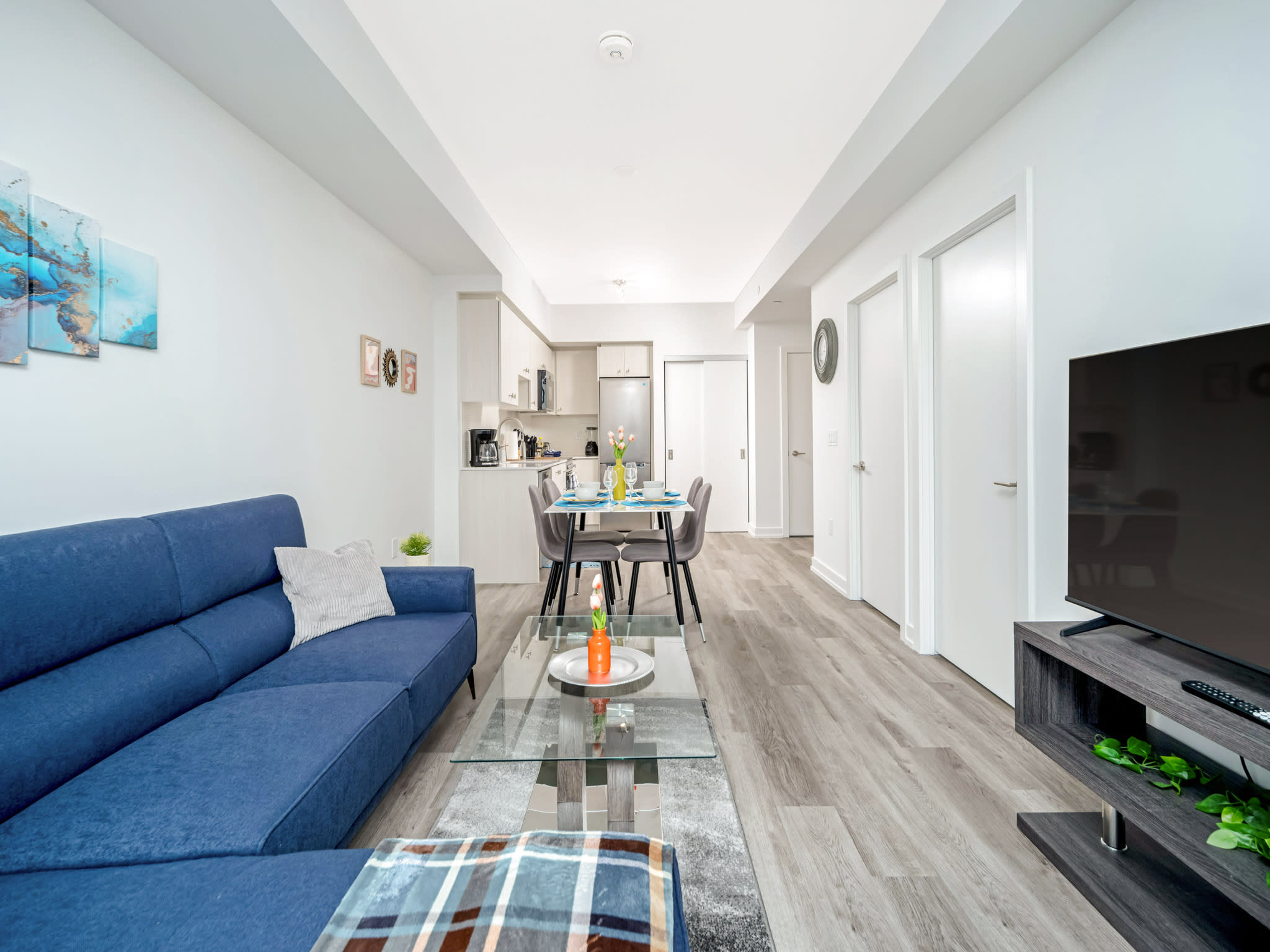 Wide shot of the open-concept living space, featuring a blue sectional sofa, glass coffee table, and TV area, extending towards the dining table and modern kitchen in the background. Light laminate flooring unifies the space.