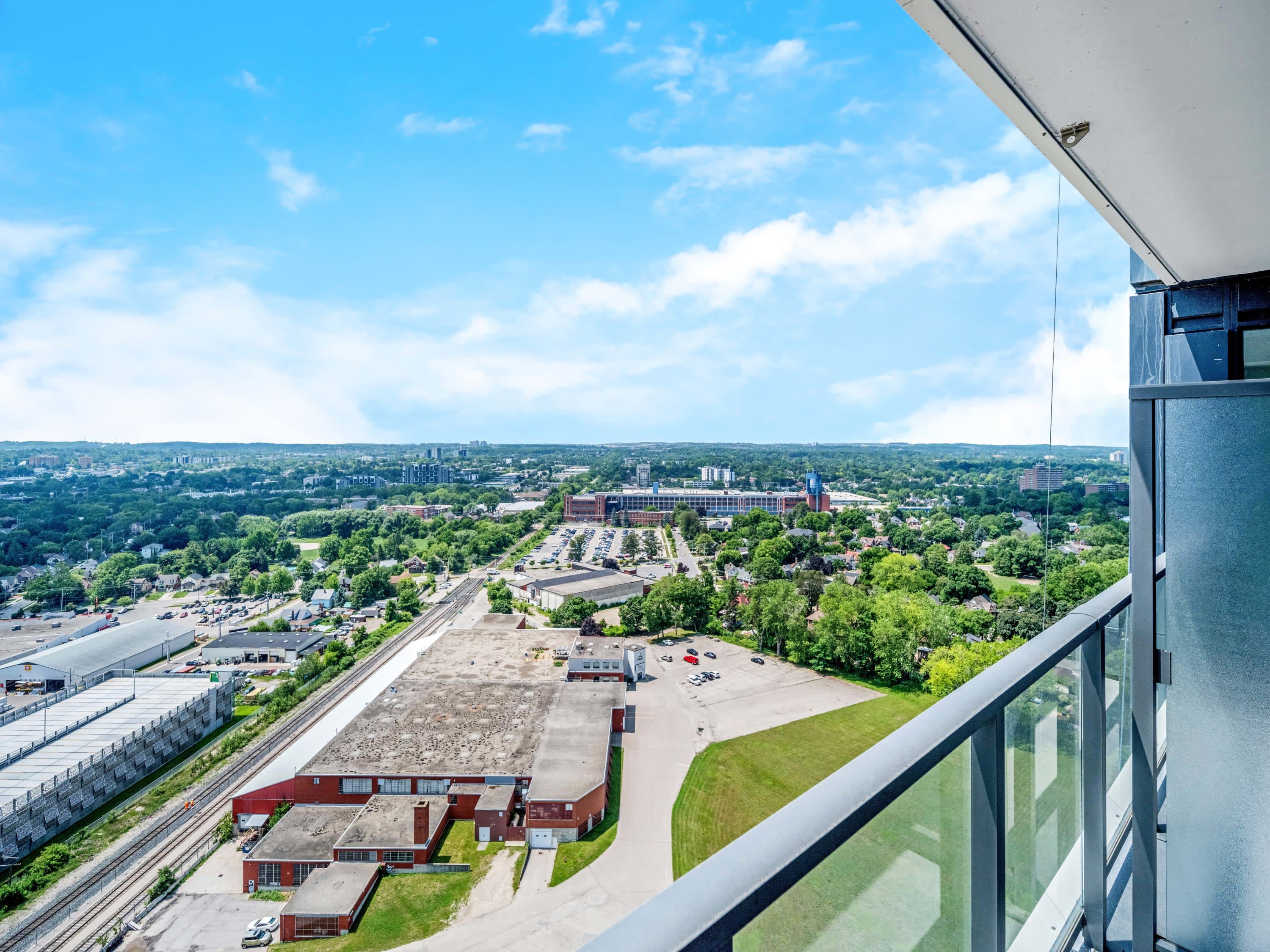 Balcony view from a high-rise, showcasing a sprawling urban landscape with industrial buildings, railway tracks, and distant residential areas under a bright blue sky. Glass railings provide an unobstructed panorama.